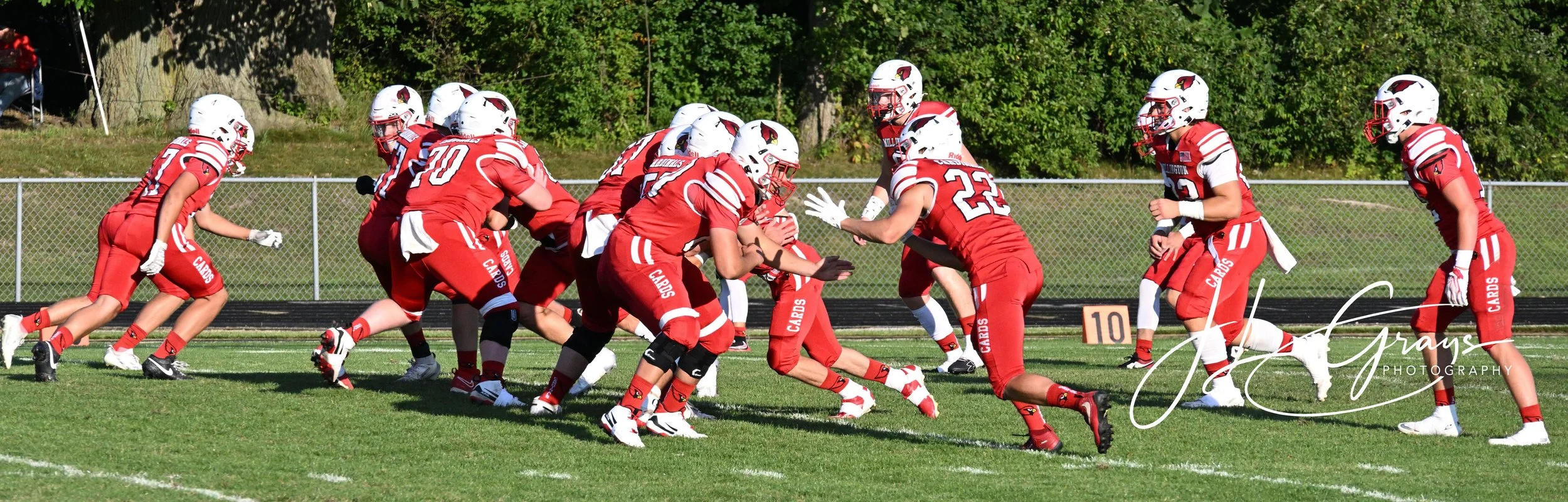A group of football players in red uniforms playing on the field, with some players blocking and others running. There is a fence and green trees in the background.