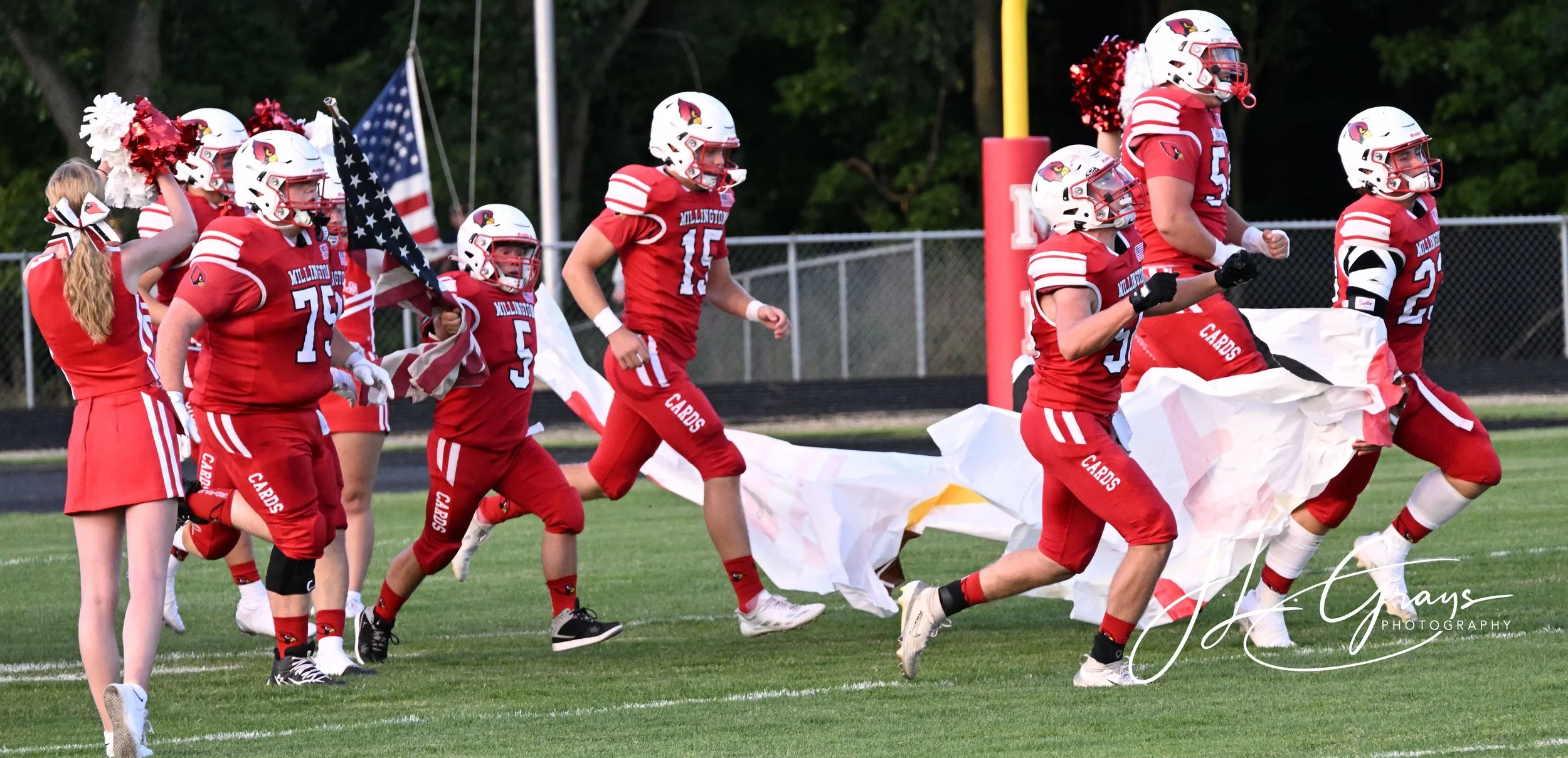 Youth football team in red uniforms running onto the field carrying a flag and a large banner, cheerleader in red holding pom-poms on the left, American flag in the background, trees, and a chain-link fence.