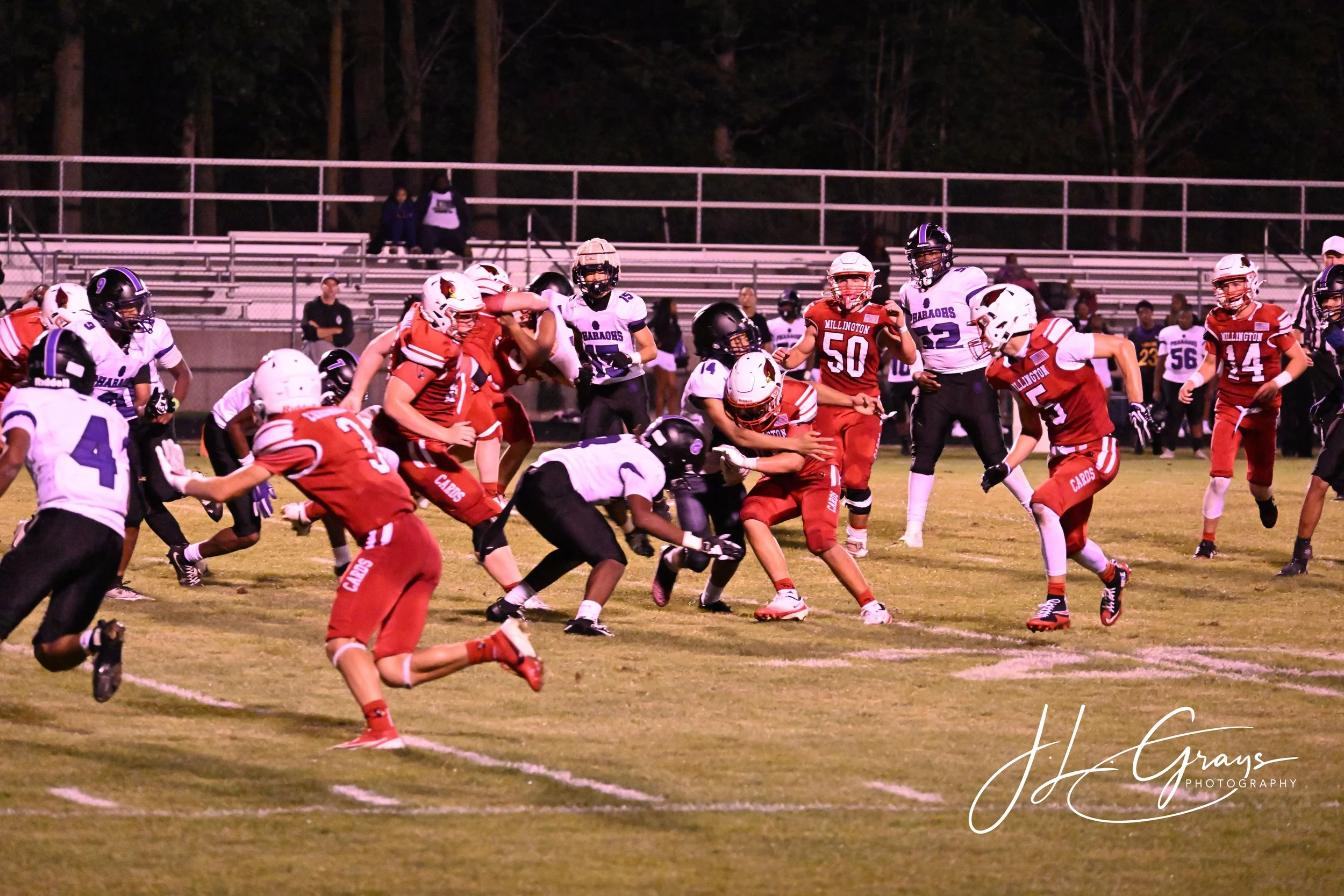 An American football game at night with players in red and white uniforms and purple and white uniforms on a grassy field. Some players are tackling while others are running or standing. Spectators sit in metal bleachers in the background, and trees 