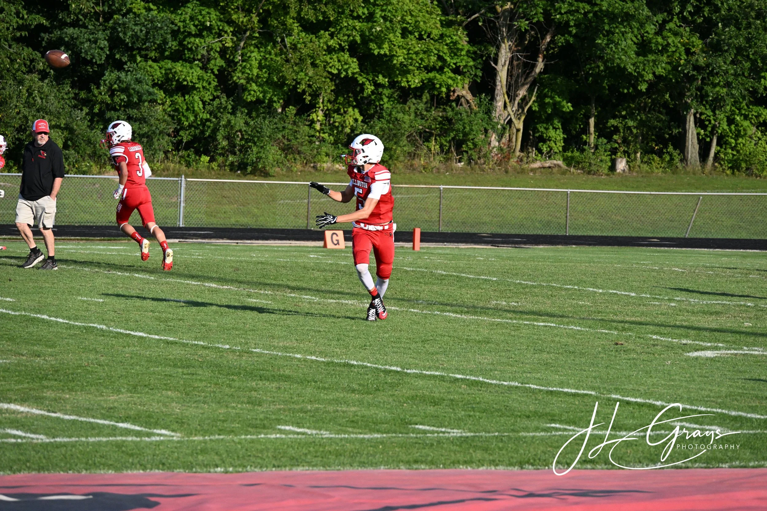 High school football players in red uniforms on the field during a game, with a coach and a lush green background.