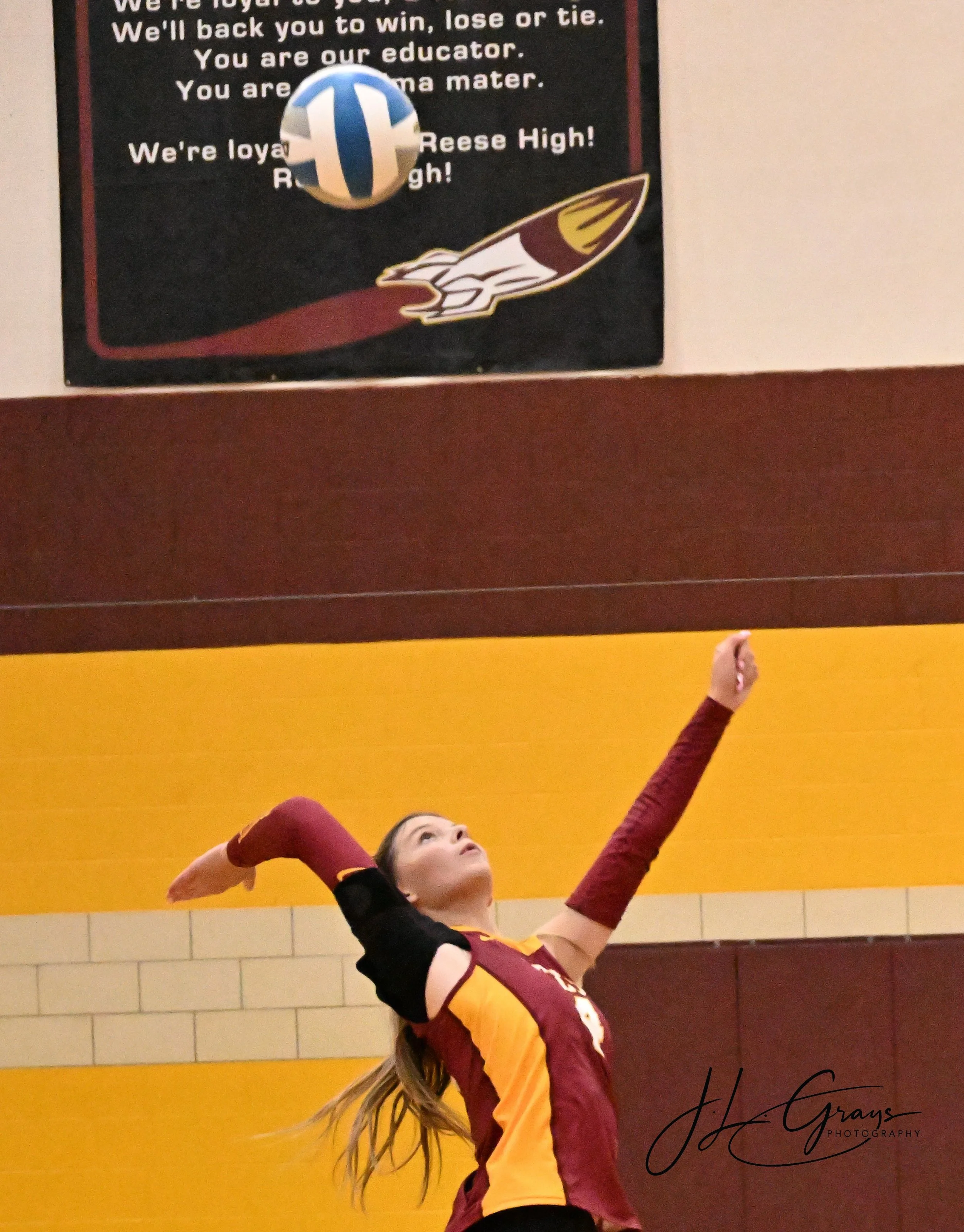 A young female volleyball player in a maroon and yellow uniform is in the middle of a jump, reaching up with her right arm to hit a volleyball against a black and yellow wall background. Her long hair is flowing down her back, and she appears focused