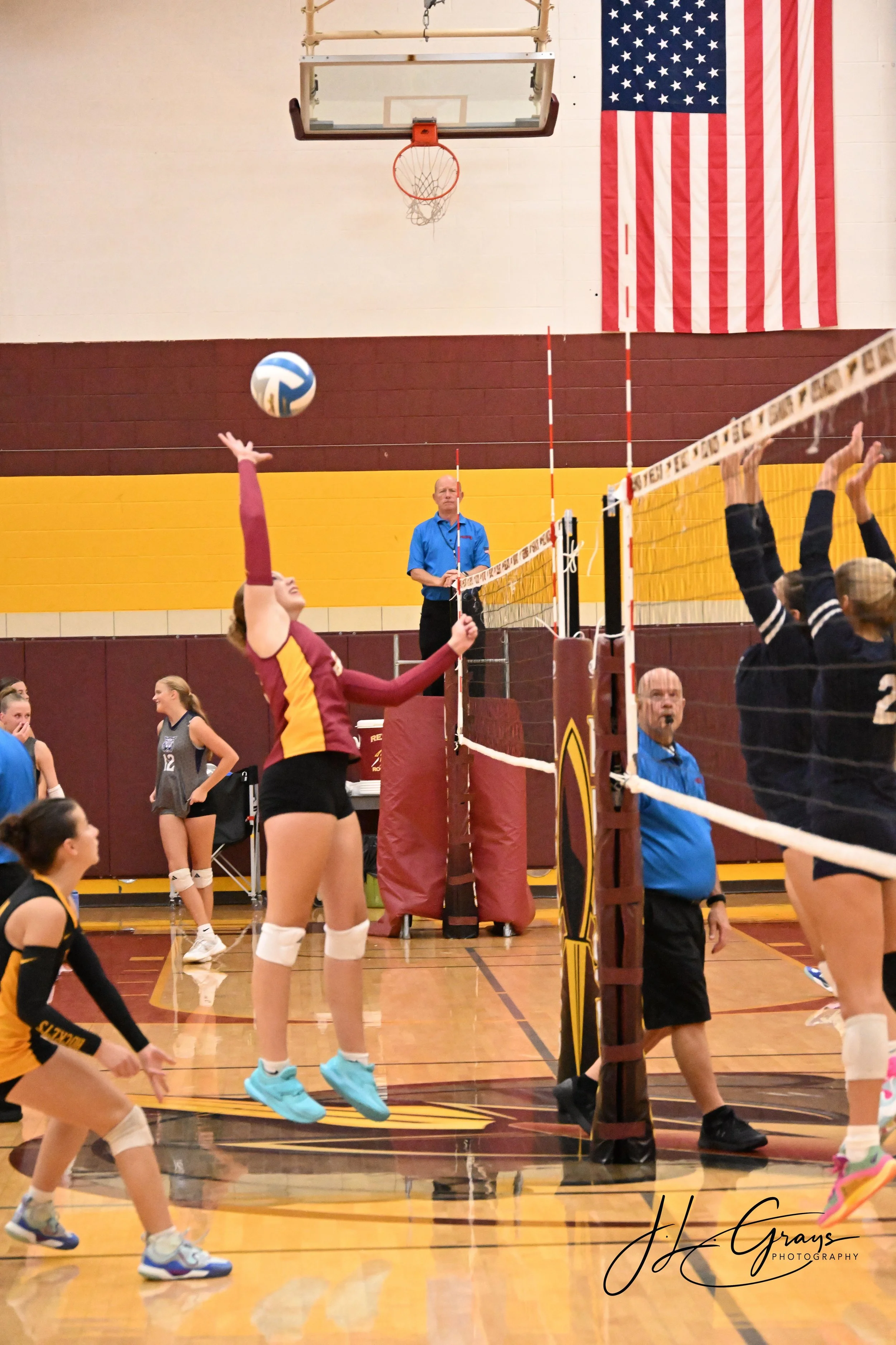 Indoor volleyball game with players in action near the net, a referee, and a large American flag in the background.