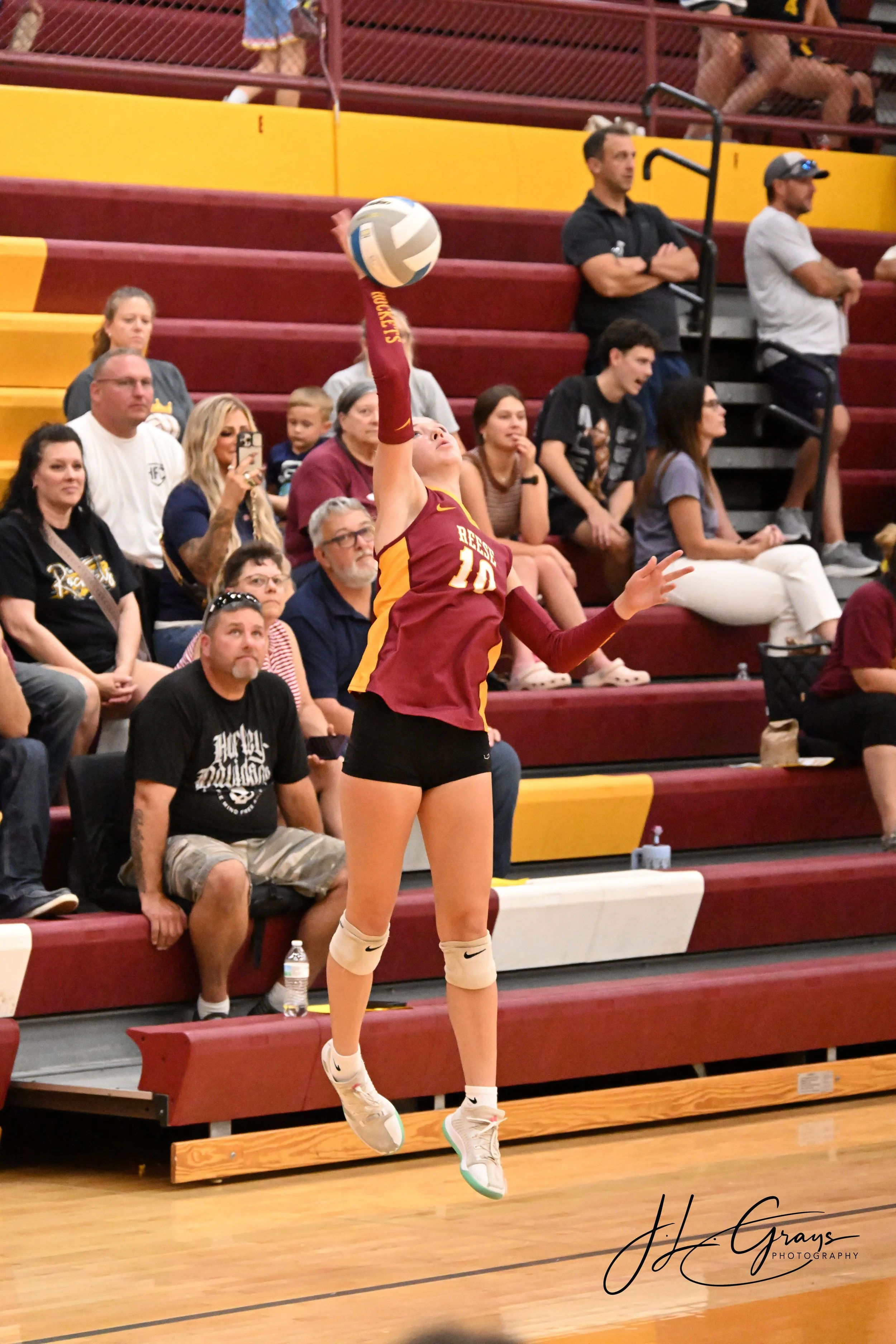 A female volleyball player in a maroon and yellow uniform with the number 10 on her jersey is jumping to hit a volleyball during a game. The scene takes place in an indoor gym with spectators sitting on bleachers, some taking photos, and others watch