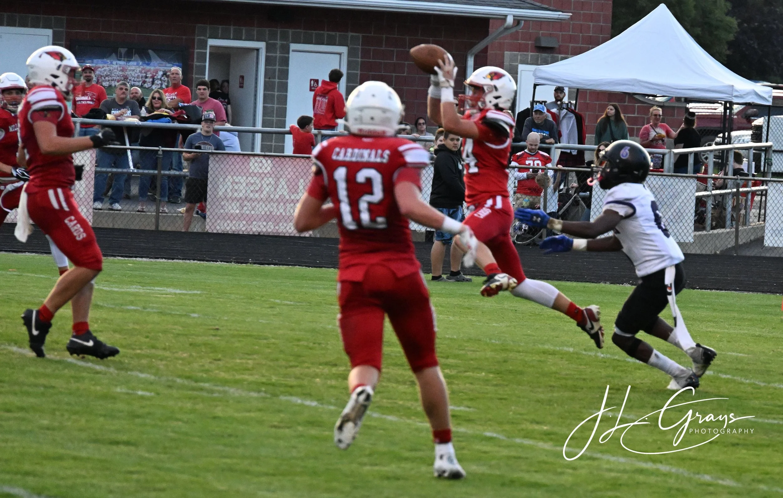 Football players in red and white uniforms playing a game on the field, with spectators watching from behind the fence.