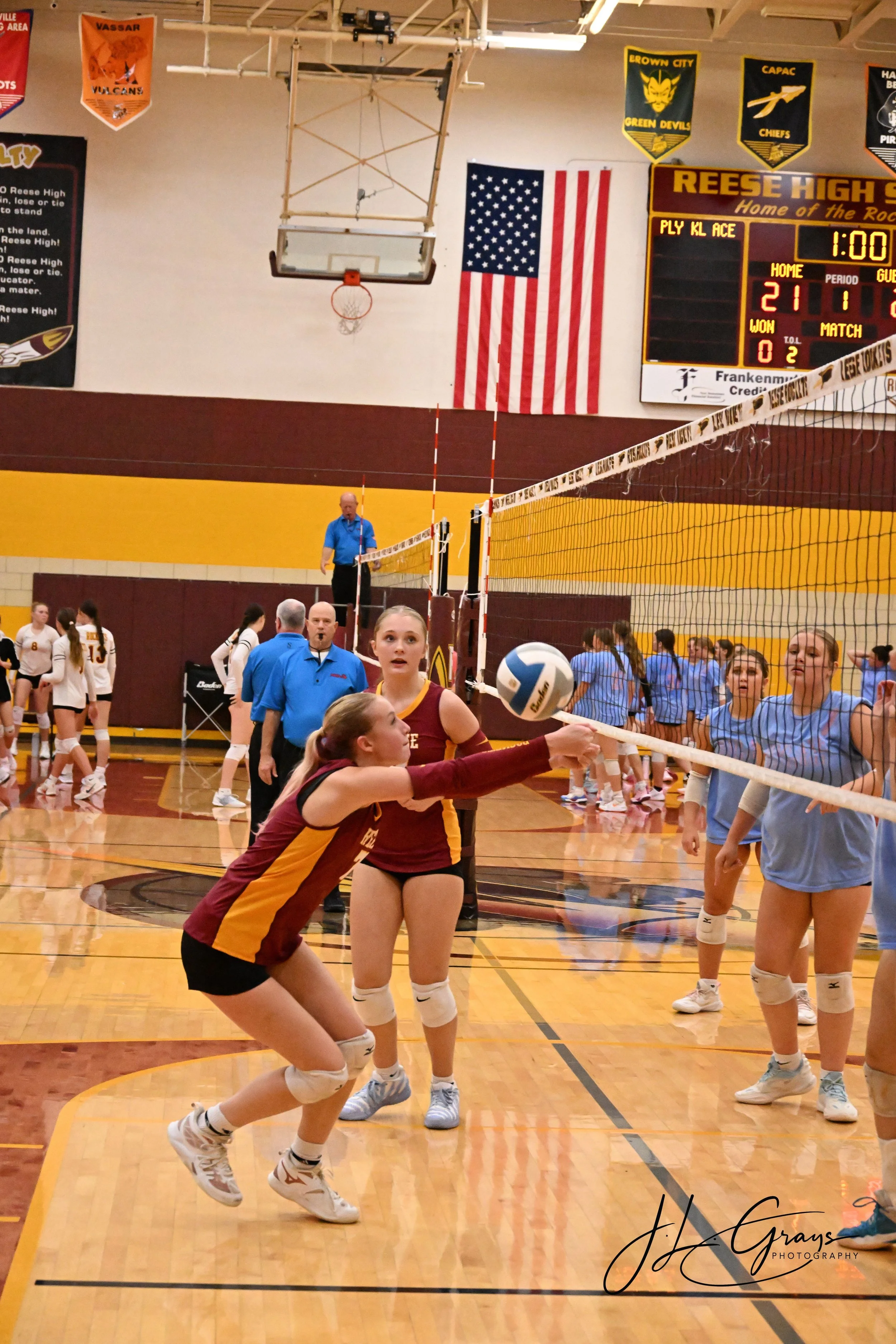 Girls playing volleyball in a gymnasium surrounded by spectators and team members, with an American flag and banners hanging on the wall.