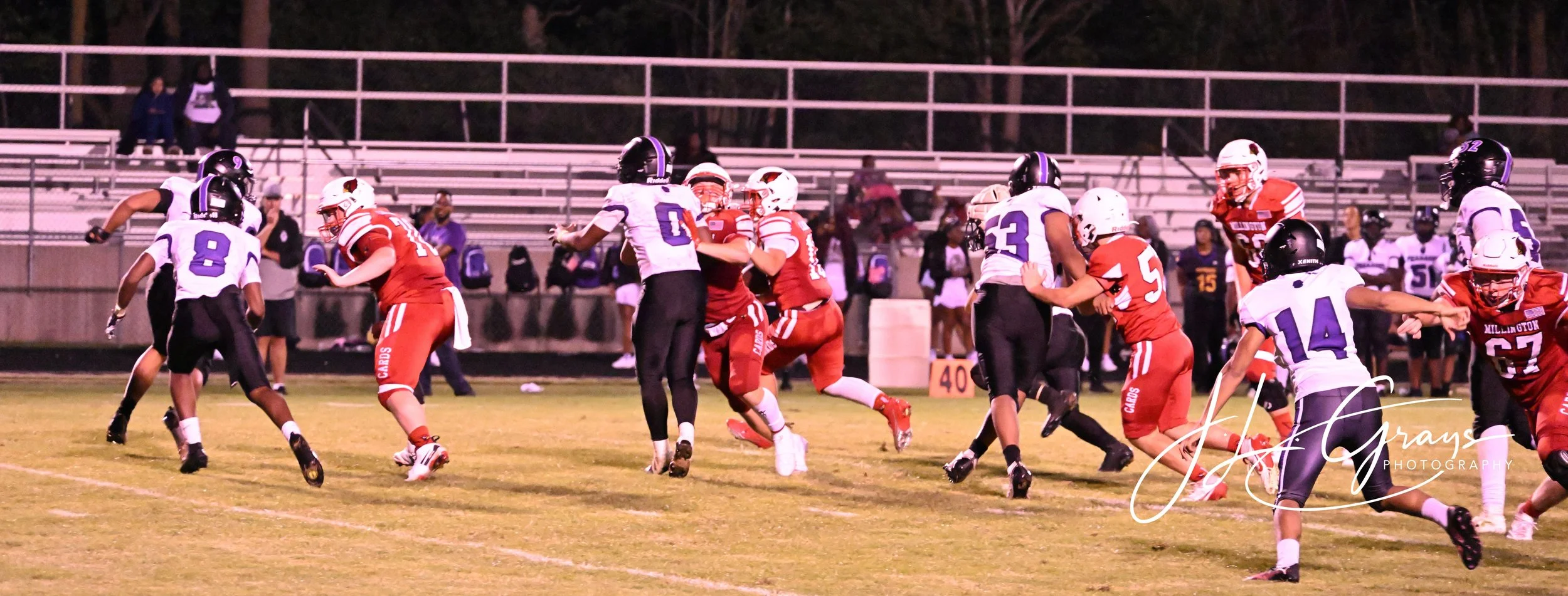 A football game in progress with players from two teams, one in white and purple uniforms and the other in red uniforms, on a grassy field under stadium lights during evening. Some players are tackling, blocking, or running with the ball, with specta