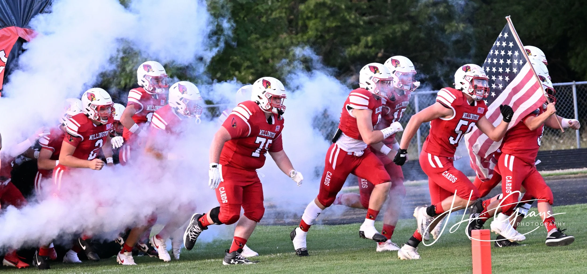Football players in red uniforms with white helmets running onto the field through smoke, carrying an American flag during a game.