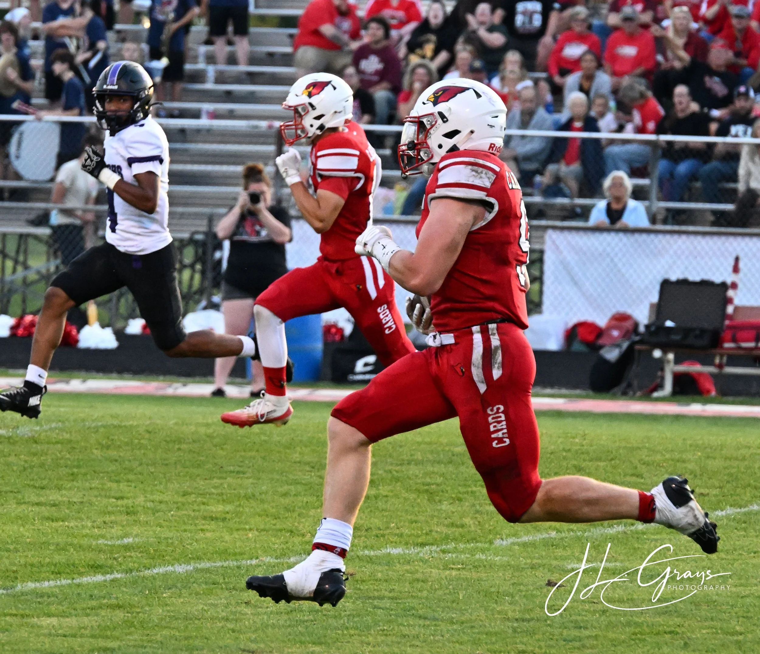 A football player in red uniform running on the field during a game, with spectators seated on bleachers in the background.