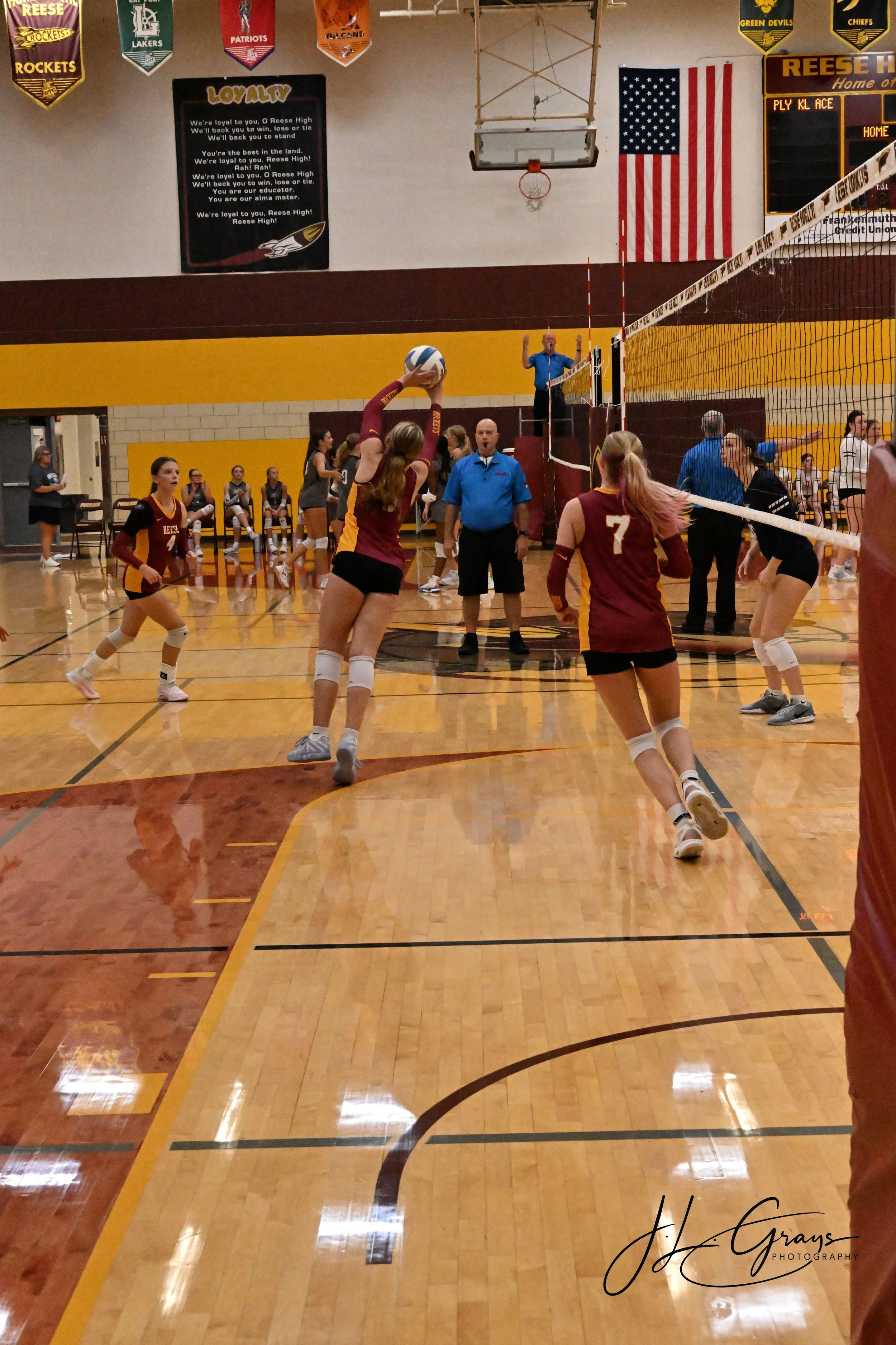 An indoor volleyball game with players on the court, a referee, and banners hanging from the gym ceiling.