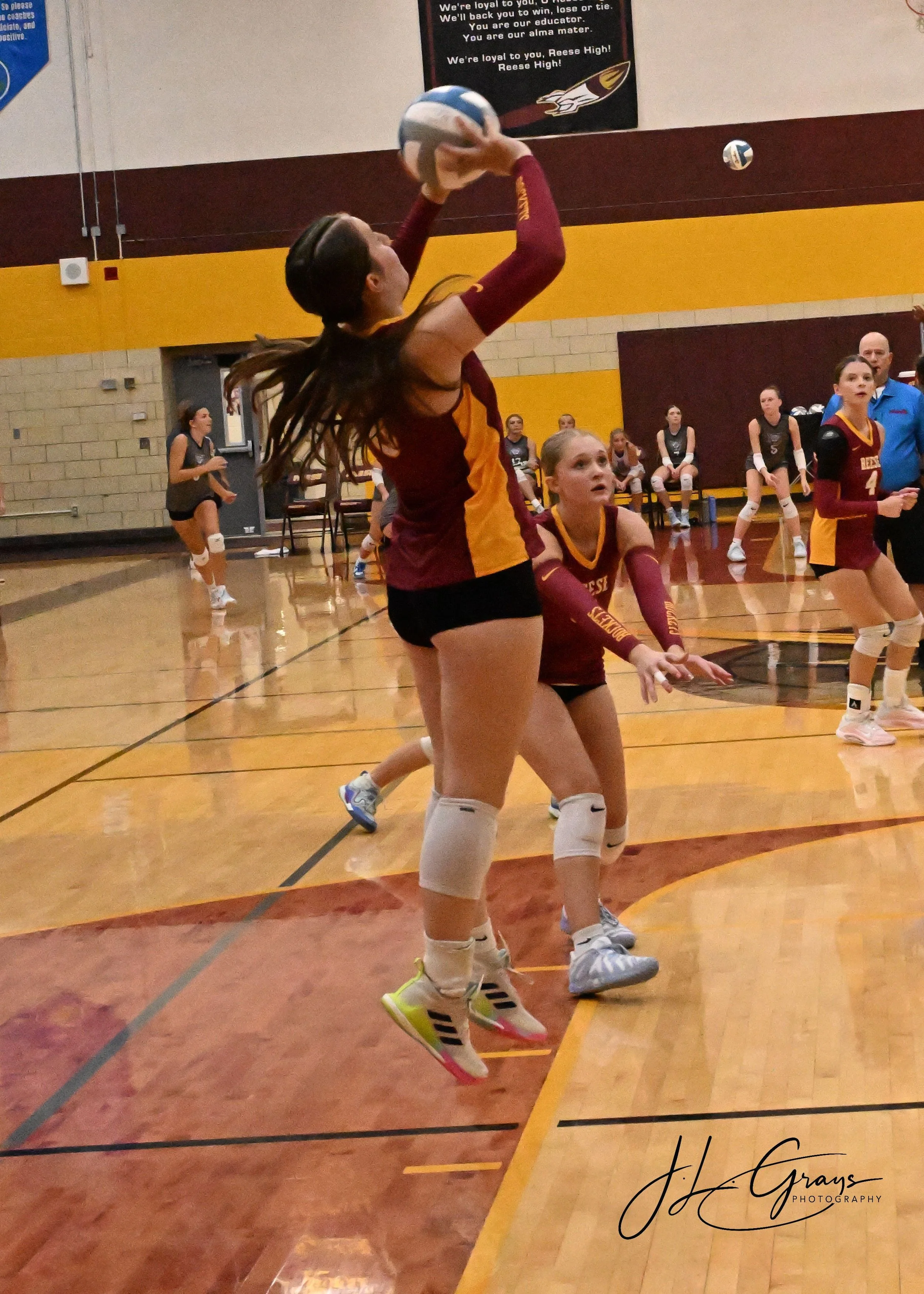 A volleyball game is underway with players in maroon and gold uniforms. One player is jumping to hit the volleyball while others are ready on the court. The background includes seated players and a coach. The setting is a gymnasium with a yellow and 