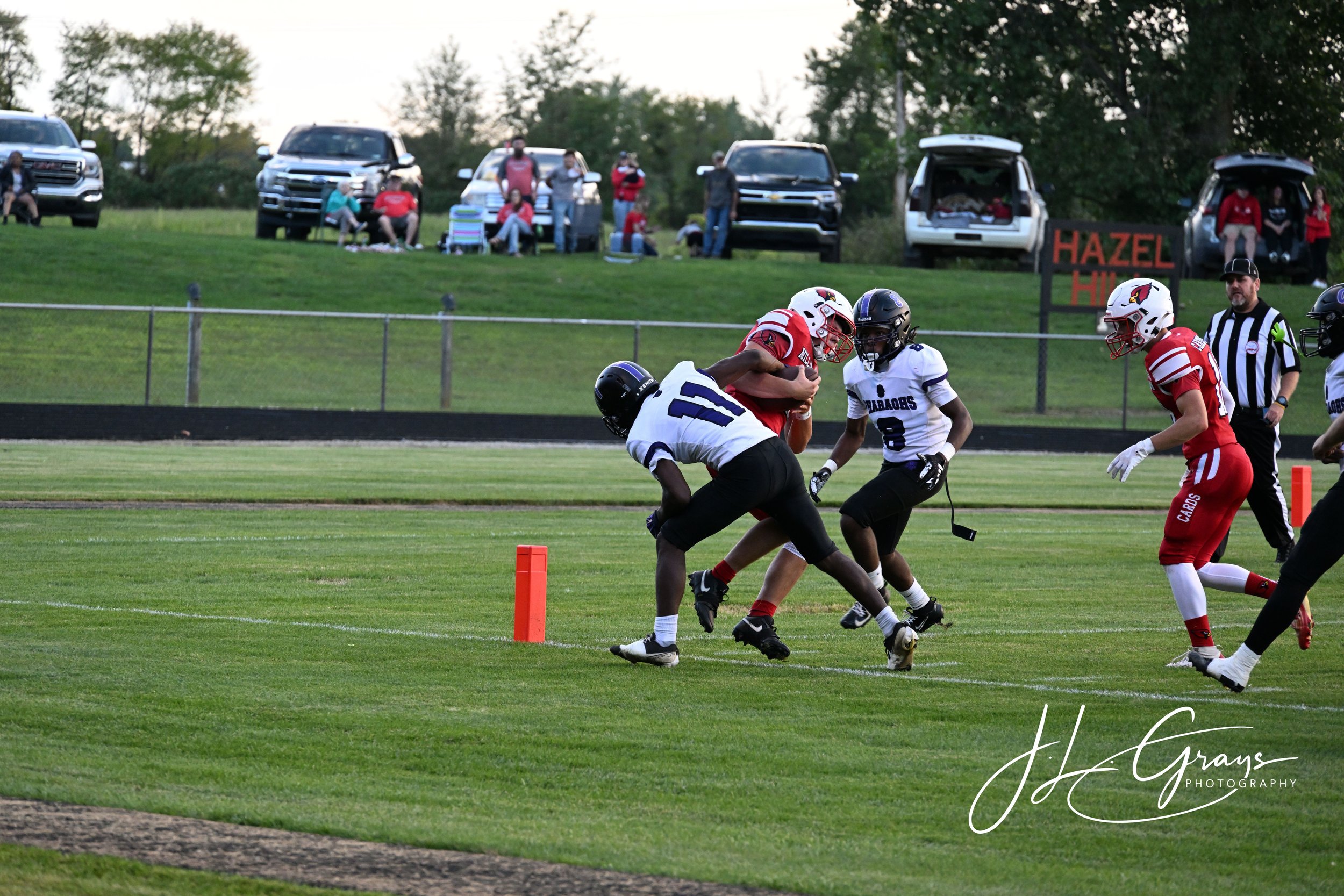 Young football players in red and white uniforms playing a game on a grassy field with spectators sitting on cars and at the top of the hill in the background.