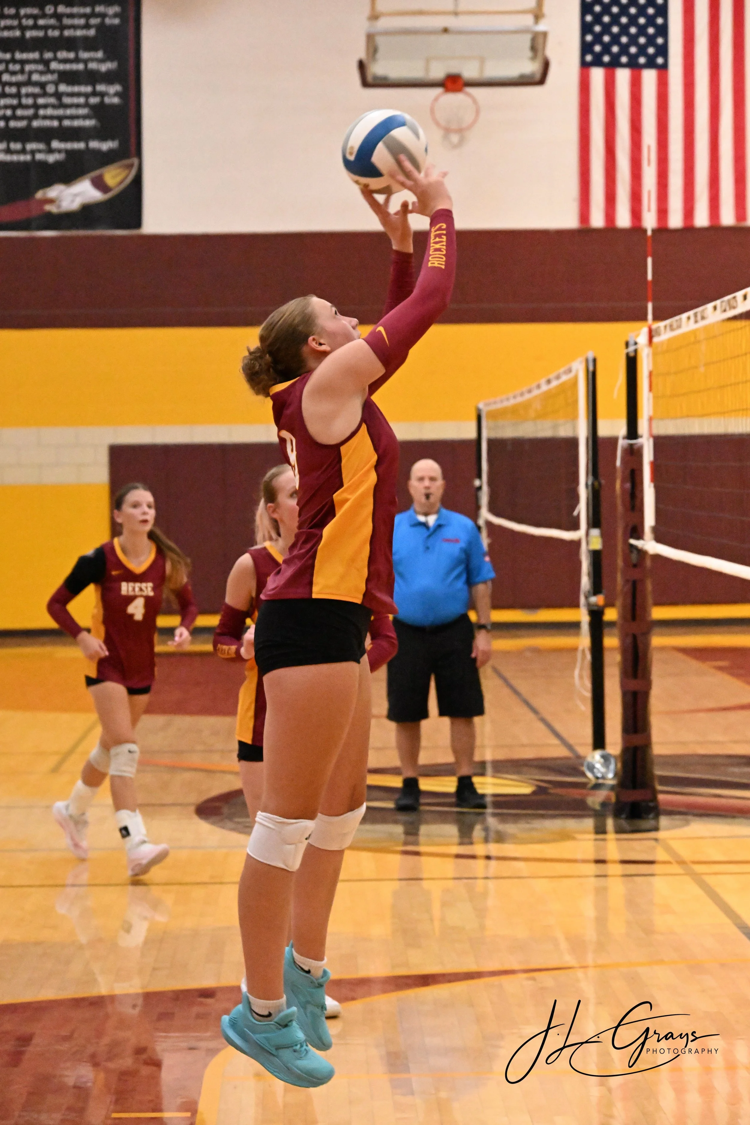 Female volleyball player wearing a maroon and gold uniform jumping to hit the ball during a game in a gym. Other players and a referee are visible in the background, with an American flag and a basketball hoop on the wall.