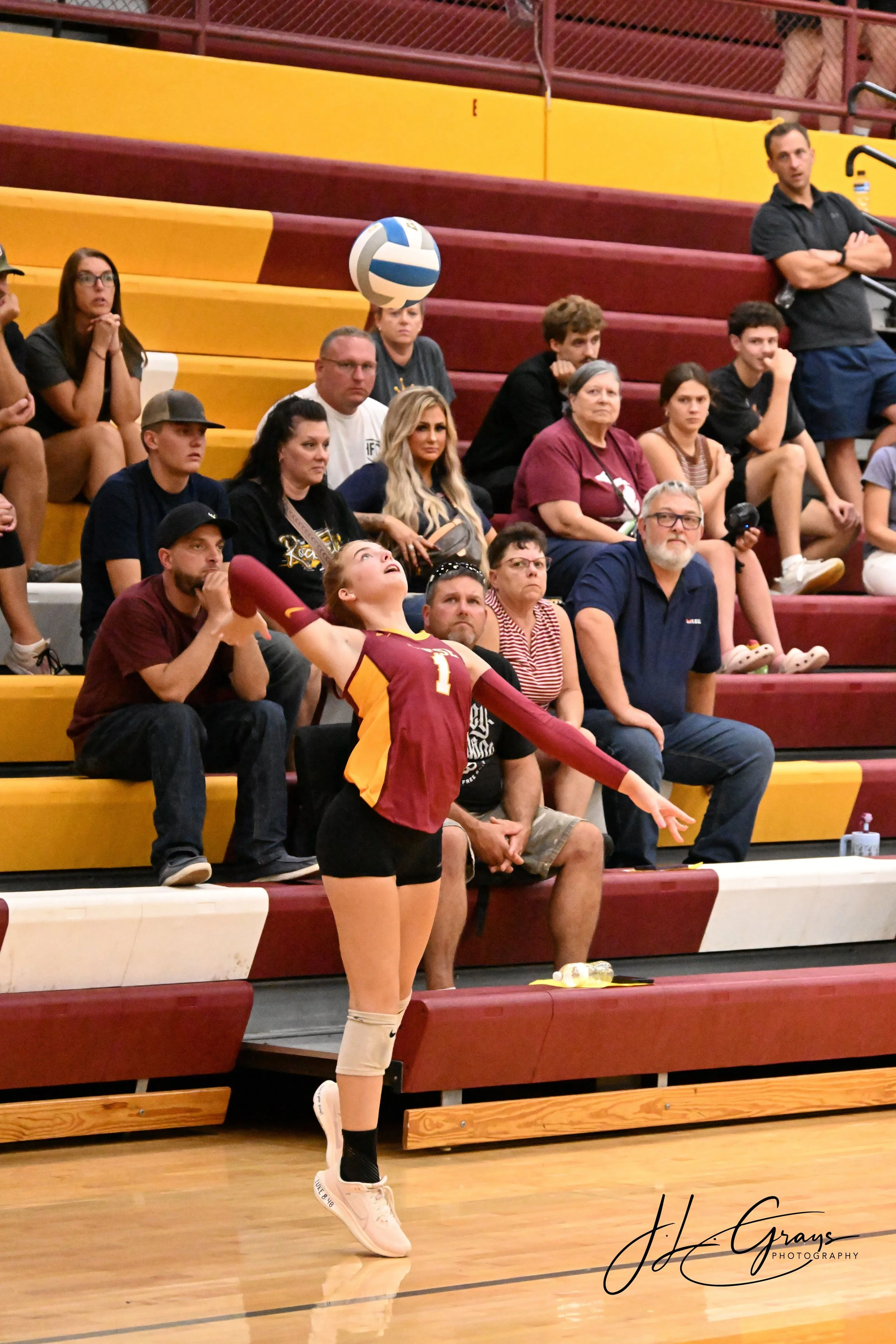 A female volleyball player in a maroon and yellow jersey wearing kneepads and sneakers is in action on an indoor court, with spectators in the background sitting on bleachers.
