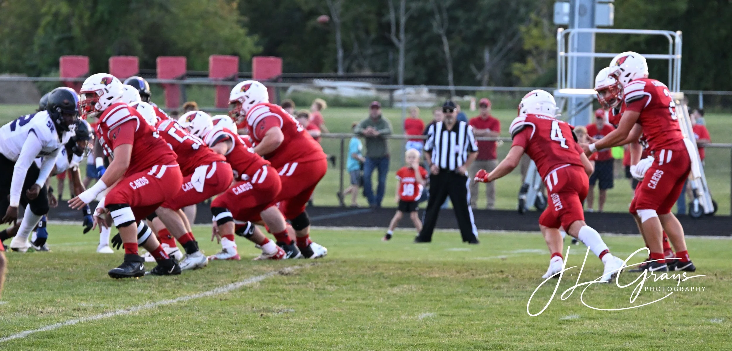 Youth football players in red uniforms and helmets line up on the field ready for a play during a game, with spectators watching in the background.