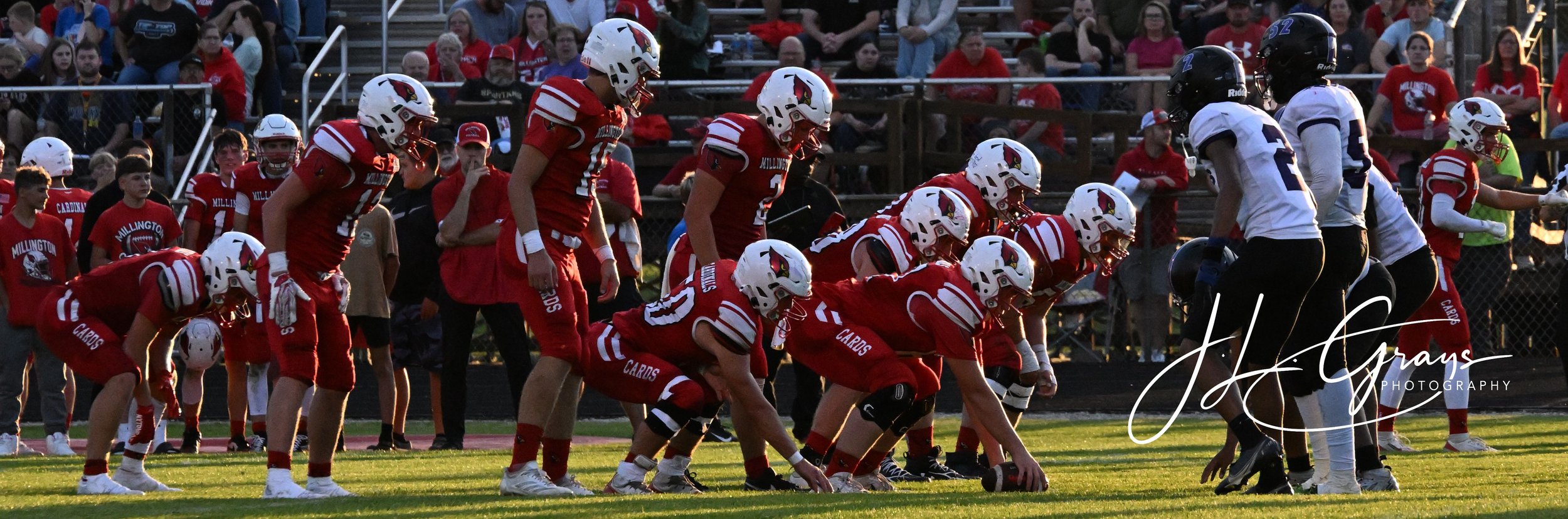 High school football game at sunset, team in red uniforms lined up at the line of scrimmage facing team in white uniforms, fans watching from bleachers in background.