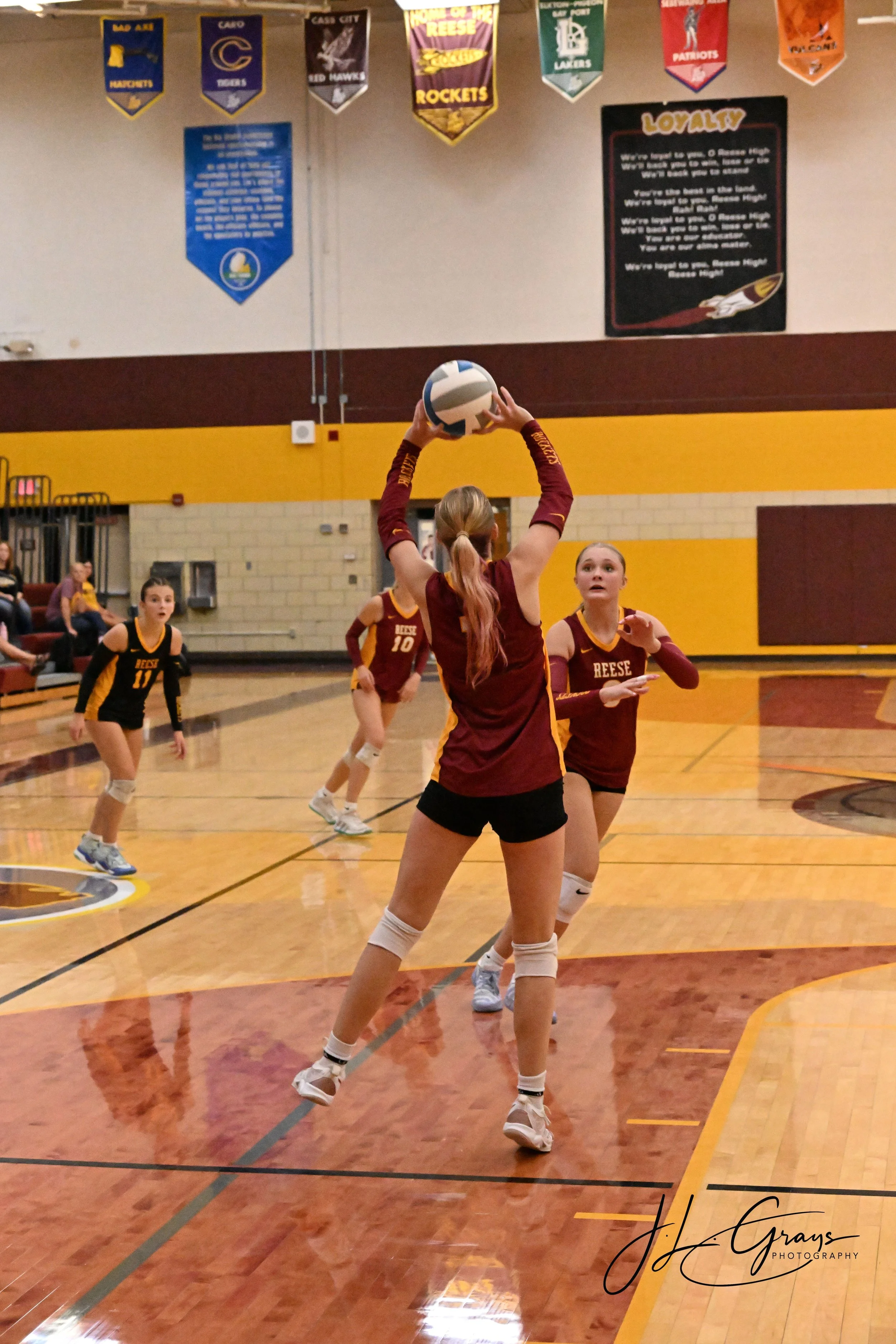 Girls playing volleyball in a gymnasium, some wearing maroon jerseys with 'Reese' written on them and others in black and yellow jerseys. One girl is jumping to hit the volleyball while others watch or prepare for the next play. Banners hang on the w