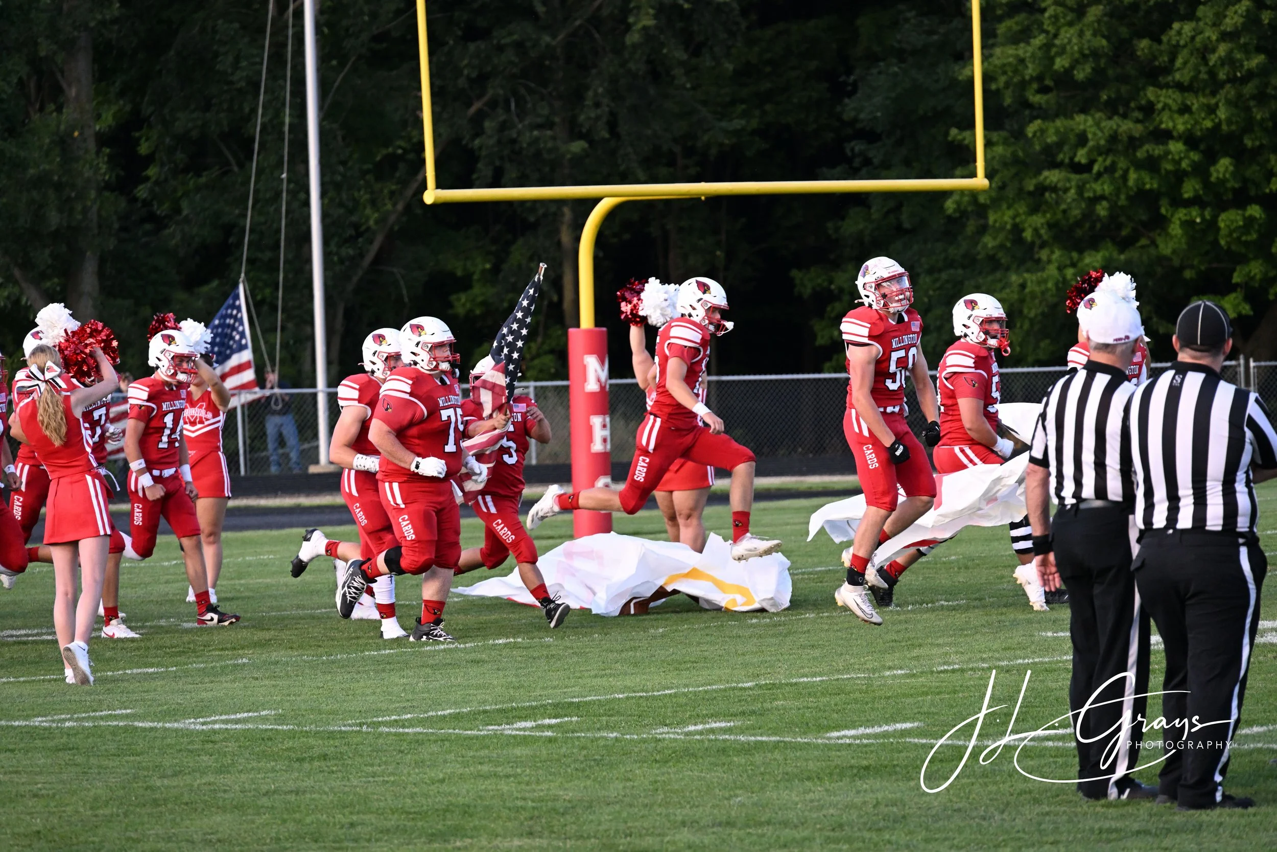 High school football players in red uniforms and white helmets running on the field while a referee nearby watches. Cheerleaders and American flags in the background with trees and a scoreboard. It appears to be during the pre-game or halftime activi