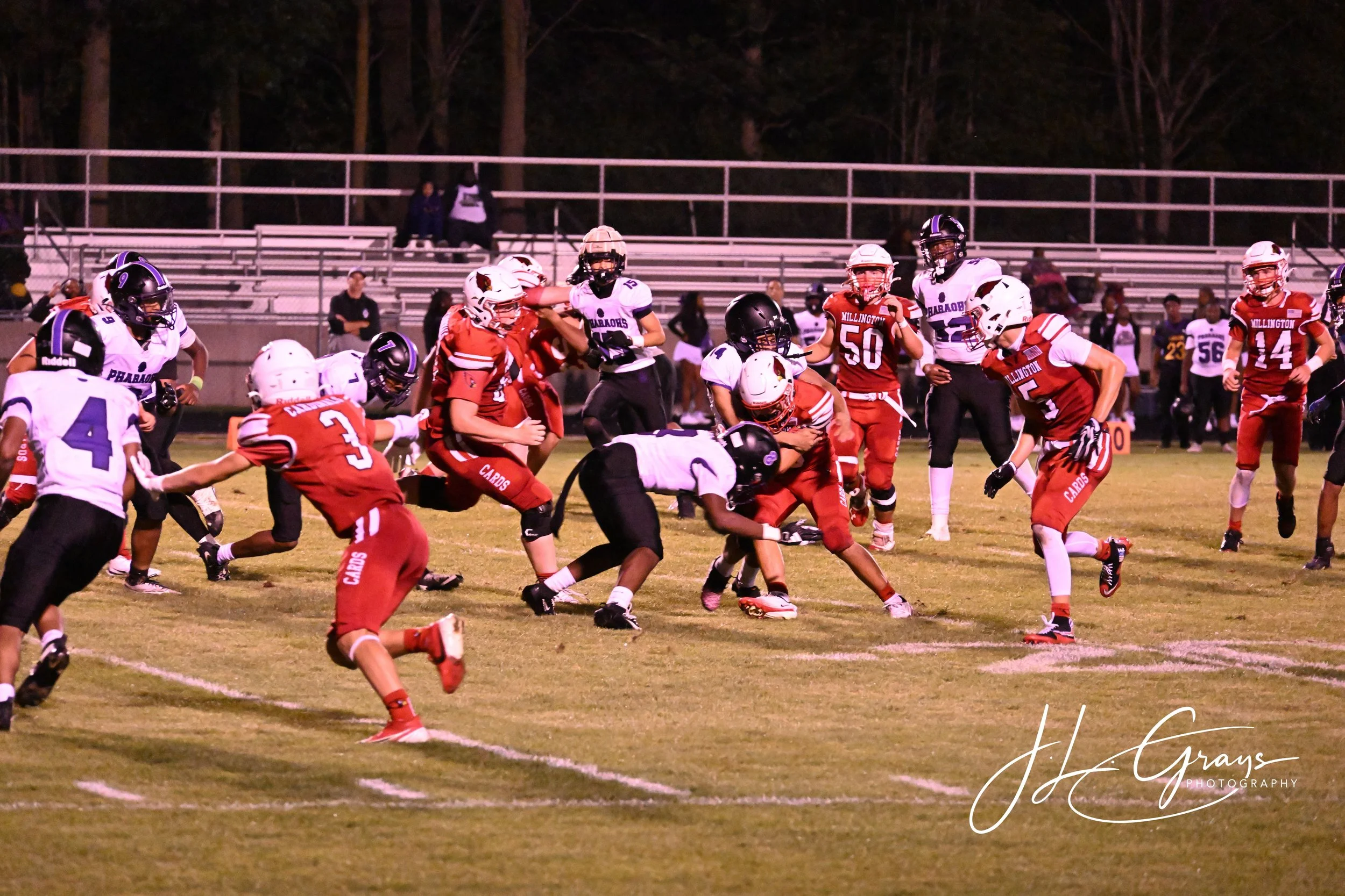 A youth football game at night, with players in red and white uniforms and players in white and purple uniforms on a grass field, with bleachers and trees in the background.