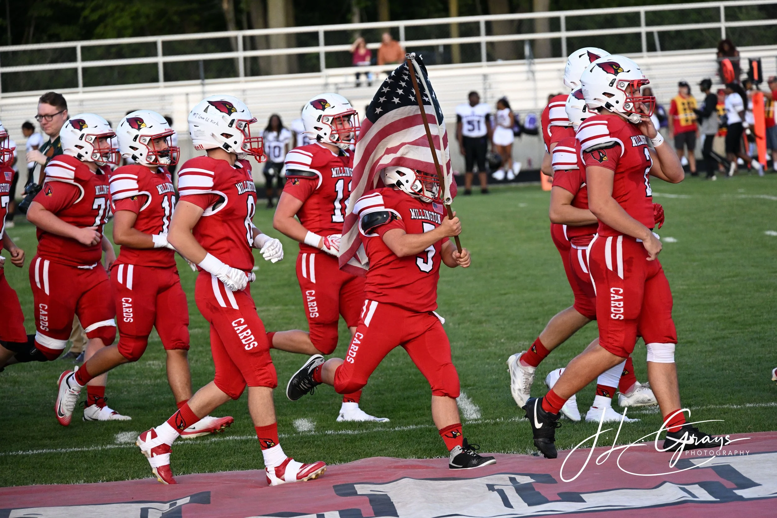High school football players in red uniforms with white helmets and red face masks walk onto the field carrying an American flag during a game. The players' uniforms have 'Millington' written on them, and the scene takes place on a football field wit