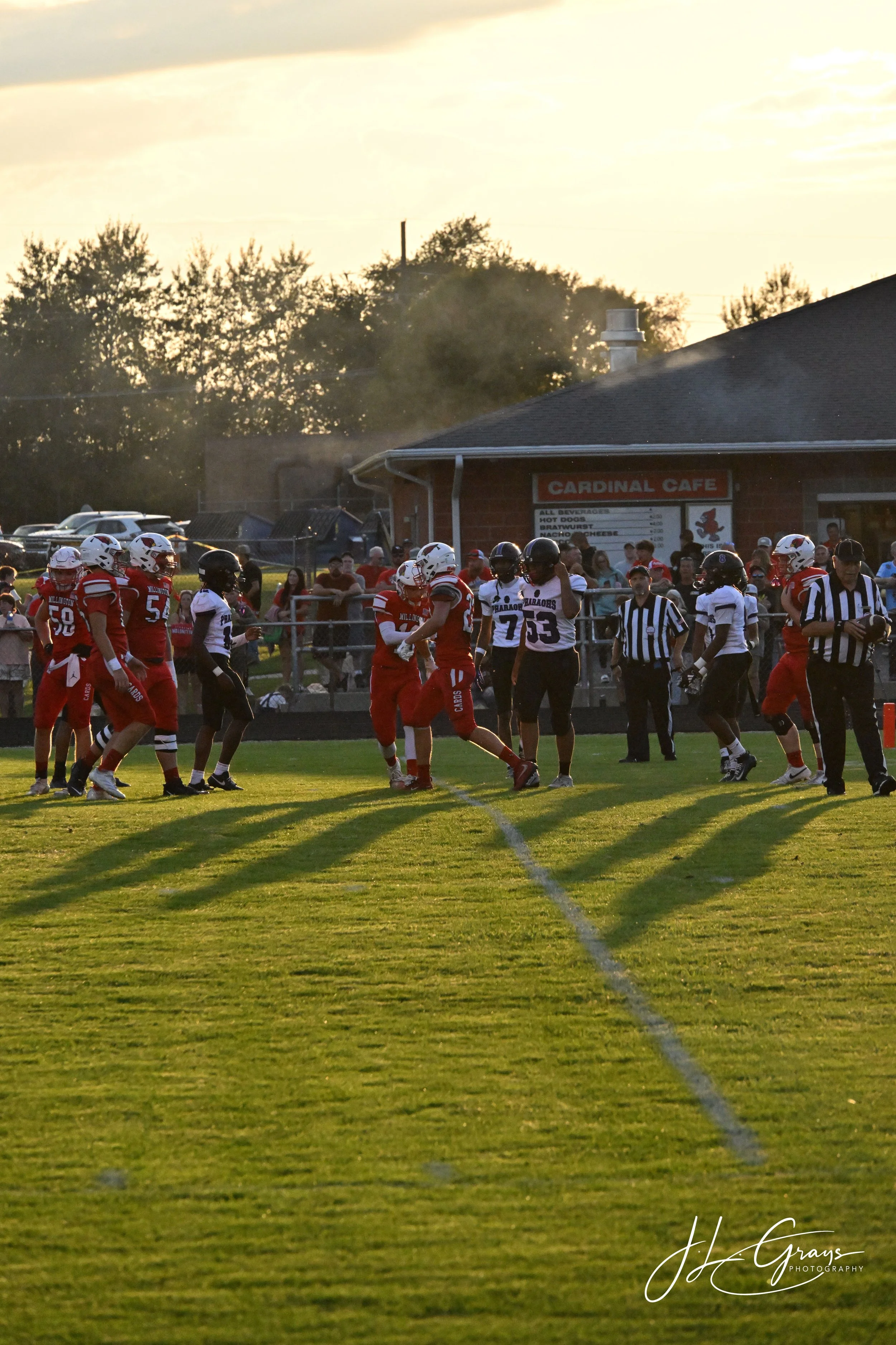 A group of football players in red and black uniforms on a grassy football field during a game, with spectators watching from the sidelines and a building with a sign for Cardinal Cafe in the background during late afternoon or early evening sunlight