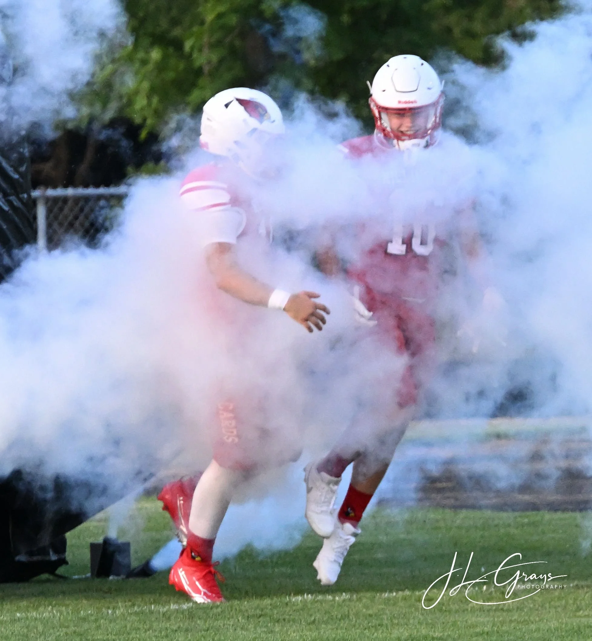 Two American football players from the same team, wearing white and red uniforms, running onto the field through smoke with a grassy field and trees in the background.