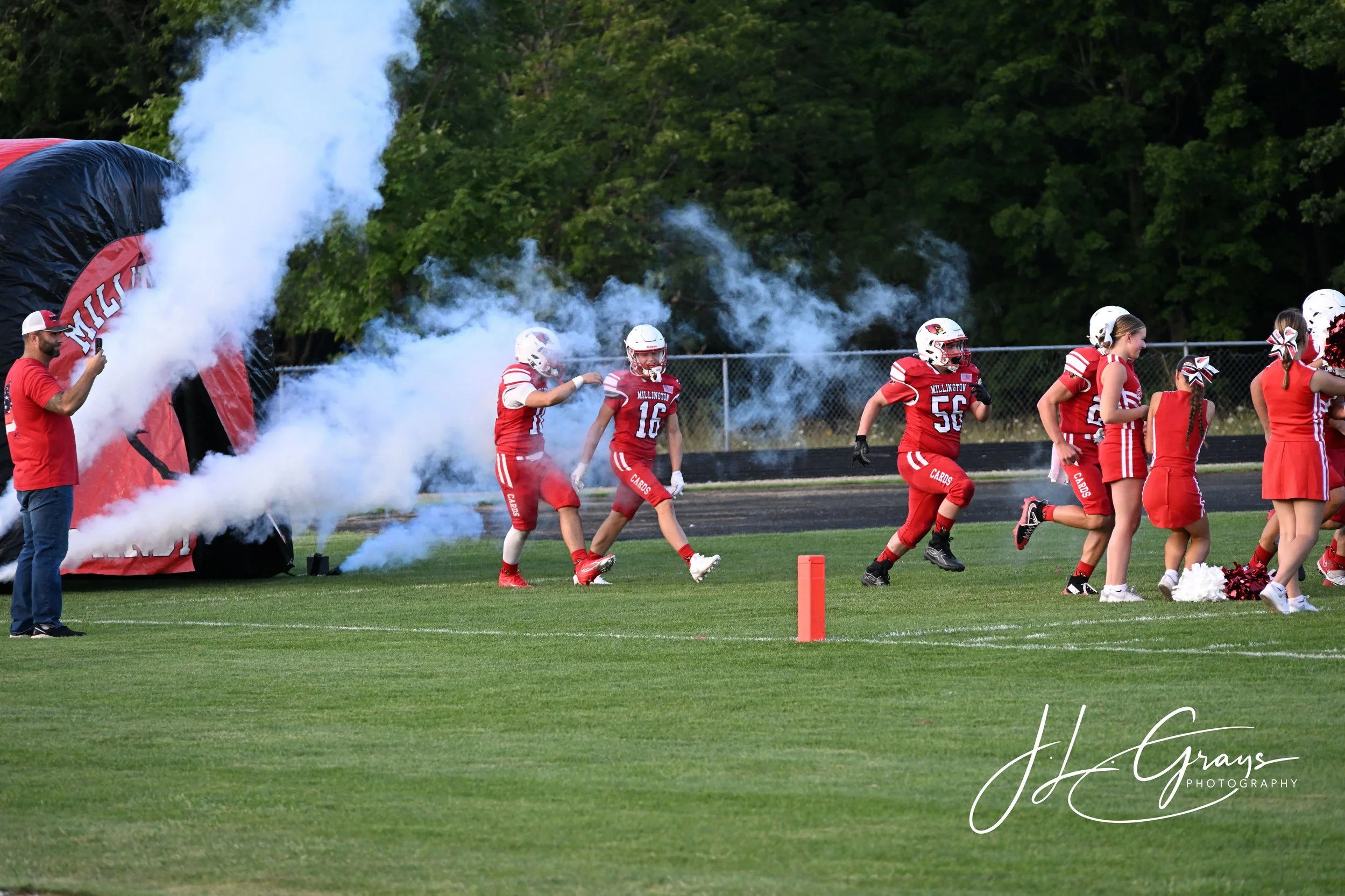 A group of young American football players in red uniforms and white helmets running onto the field through a cloud of smoke during a game, with trees in the background.
