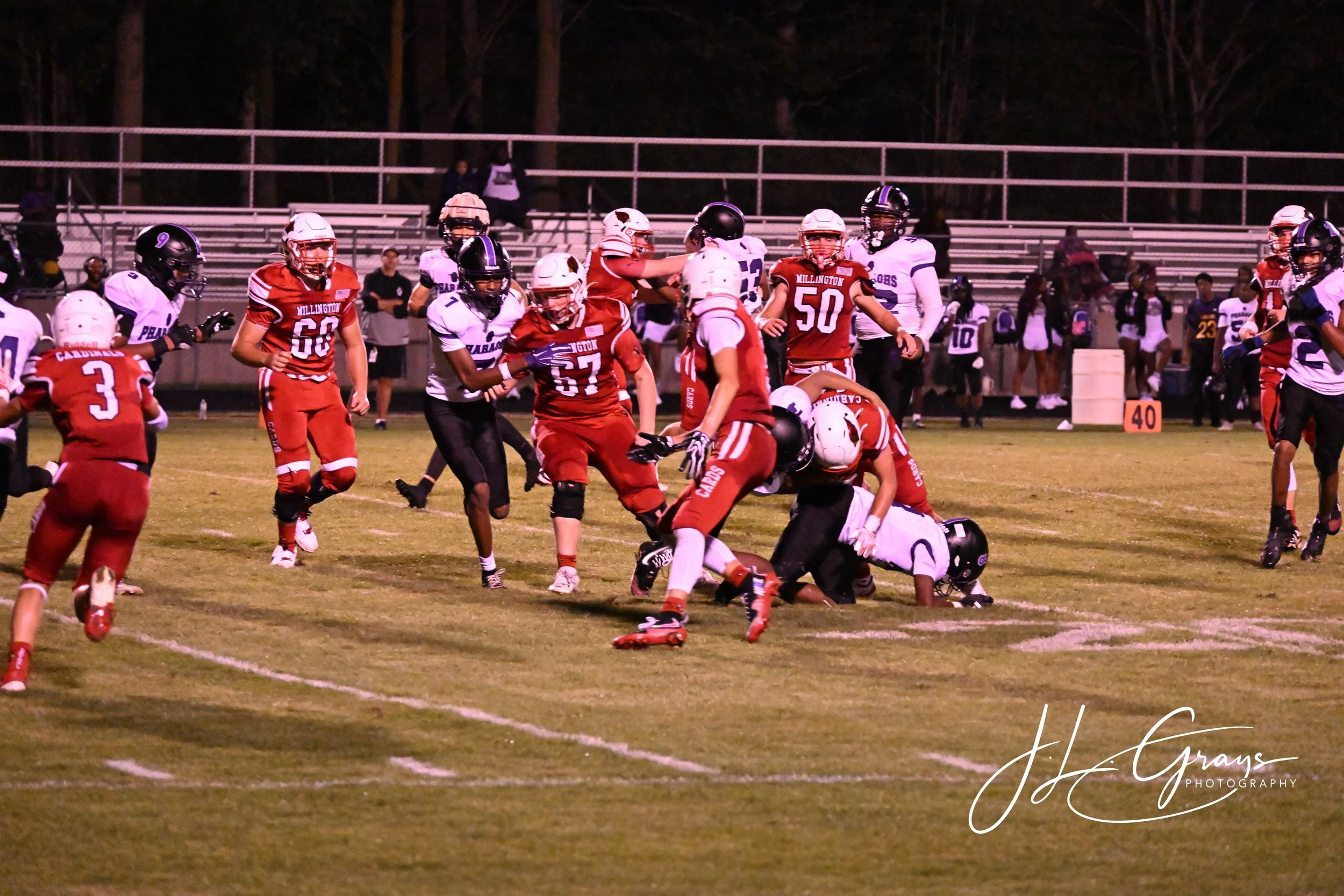 An American football game at night with players in red and white uniforms on the field. The players are engaged in a struggle for possession of the football, with some on the ground and others standing. Bleachers and spectators are visible in the bac