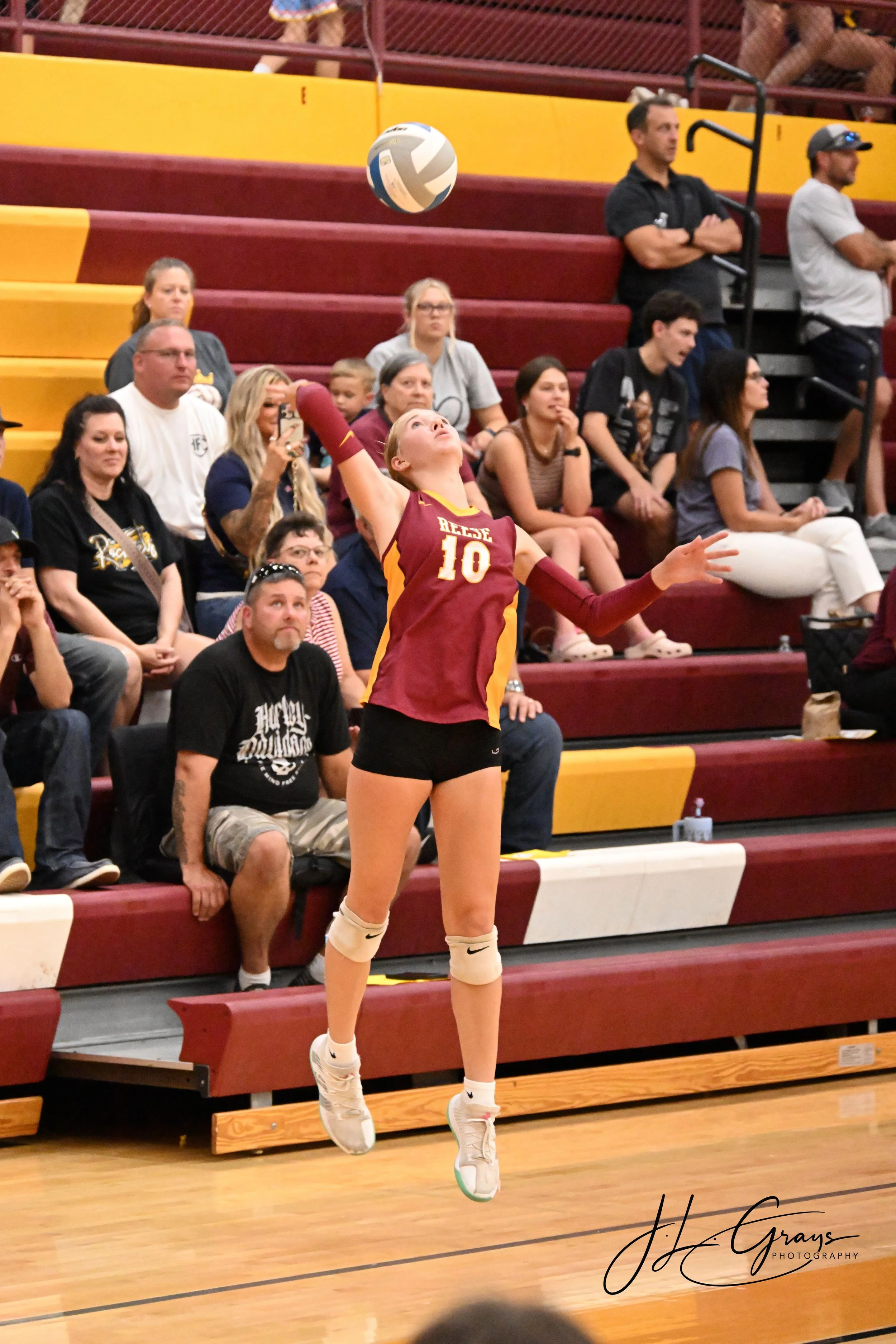 A female volleyball player in a maroon and yellow jersey with the number 10 in midair, preparing to hit a volleyball inside a gymnasium with spectators seated on yellow and maroon bleachers.