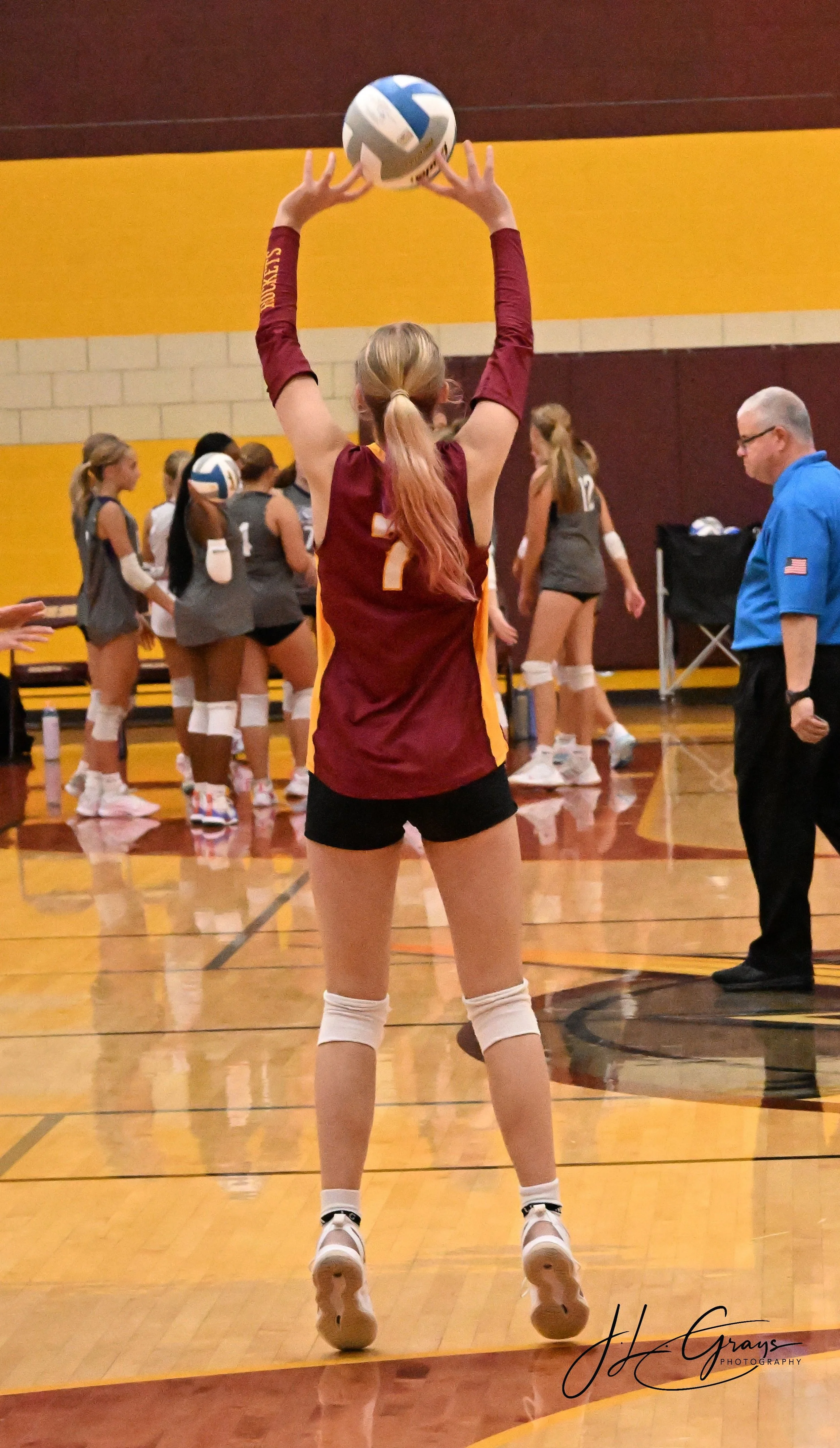 A volleyball player in a maroon and yellow jersey preparing to serve the ball on an indoor court, with teammates and a referee in the background.