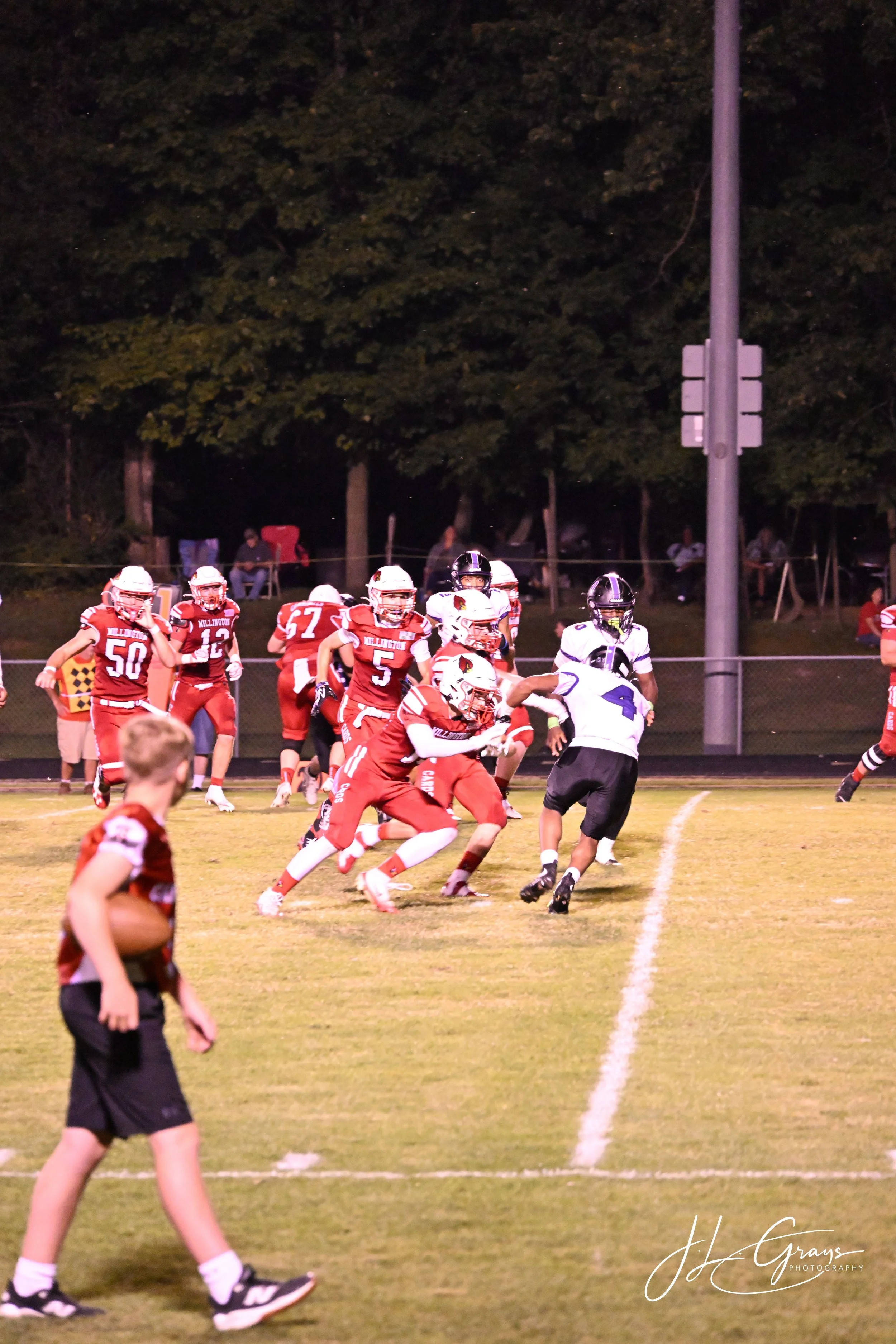 A youth football game at night, with players in red and white uniforms on the field, some running and some standing, with a referee in black uniform and a background of trees and spectators.