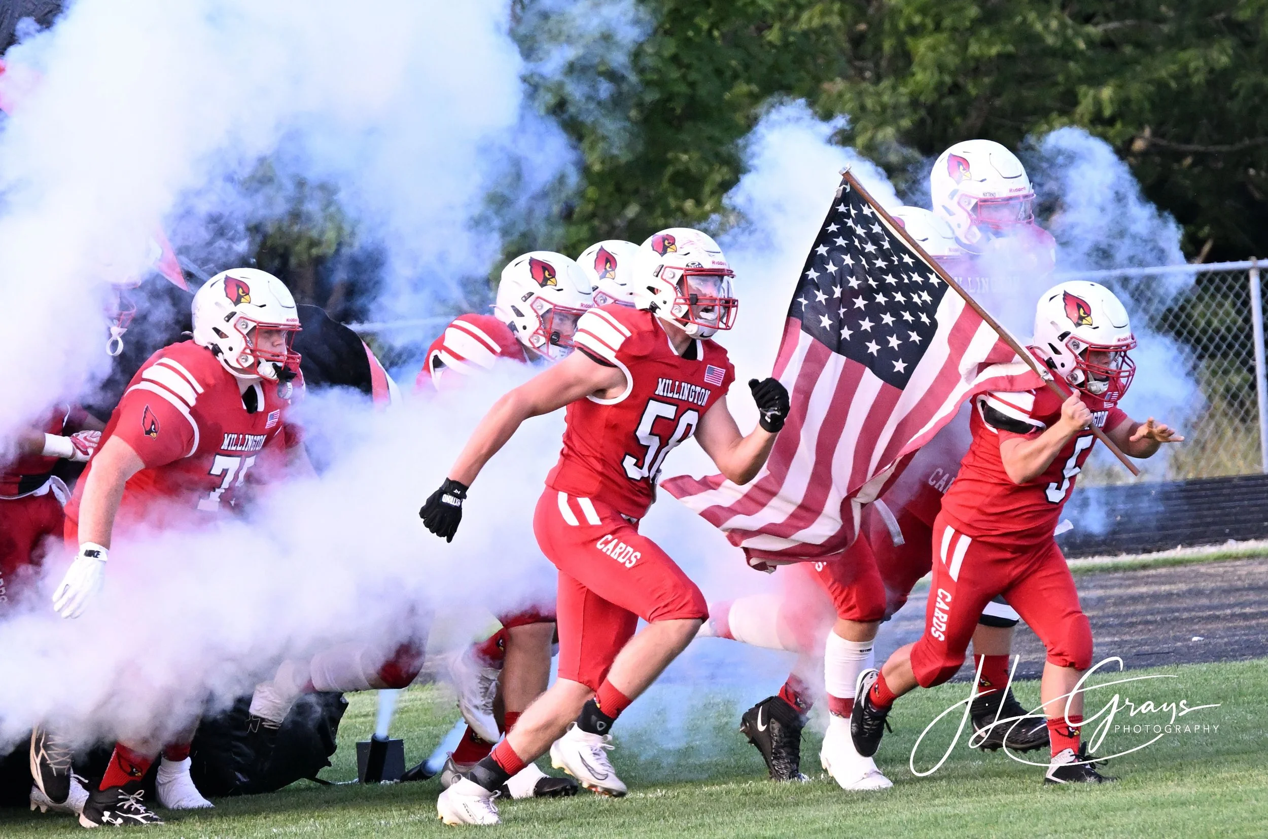 A group of football players wearing red jerseys and white helmets with a cardinal logo, running onto the field through smoke, with one player carrying an American flag.