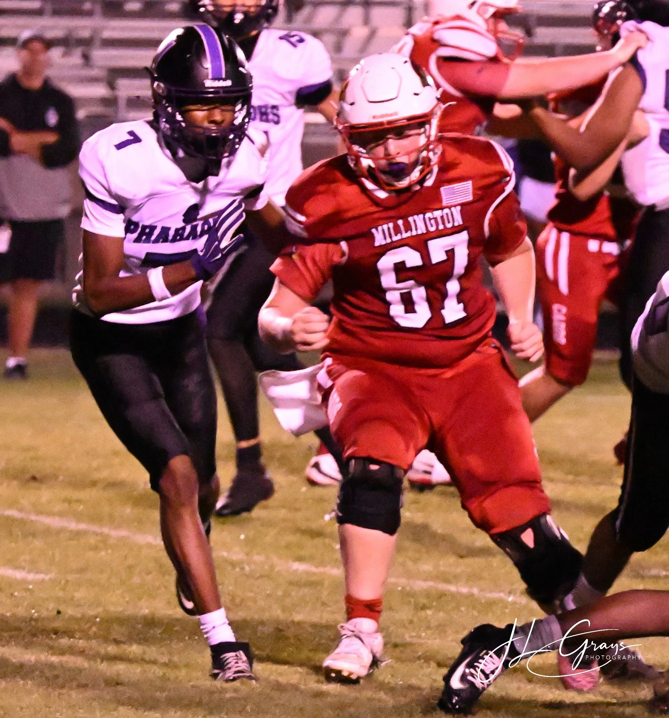 A group of high school football players on the field during a game. The players are wearing helmets, uniforms, and football gear, with some in red and others in white. They appear to be in motion, possibly running or preparing for a play. Spectators 