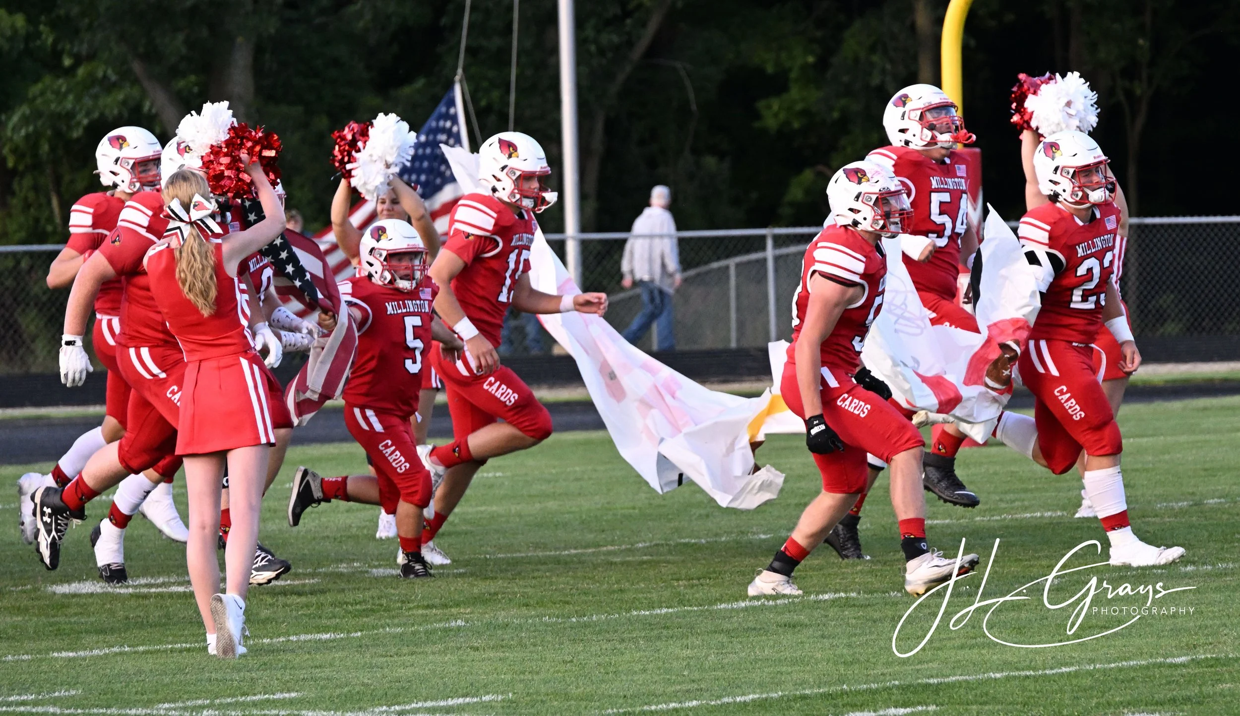 Football team in red uniforms and helmets running onto the field, some players holding cheerleading pom-poms and flags, at sunset with trees in the background.