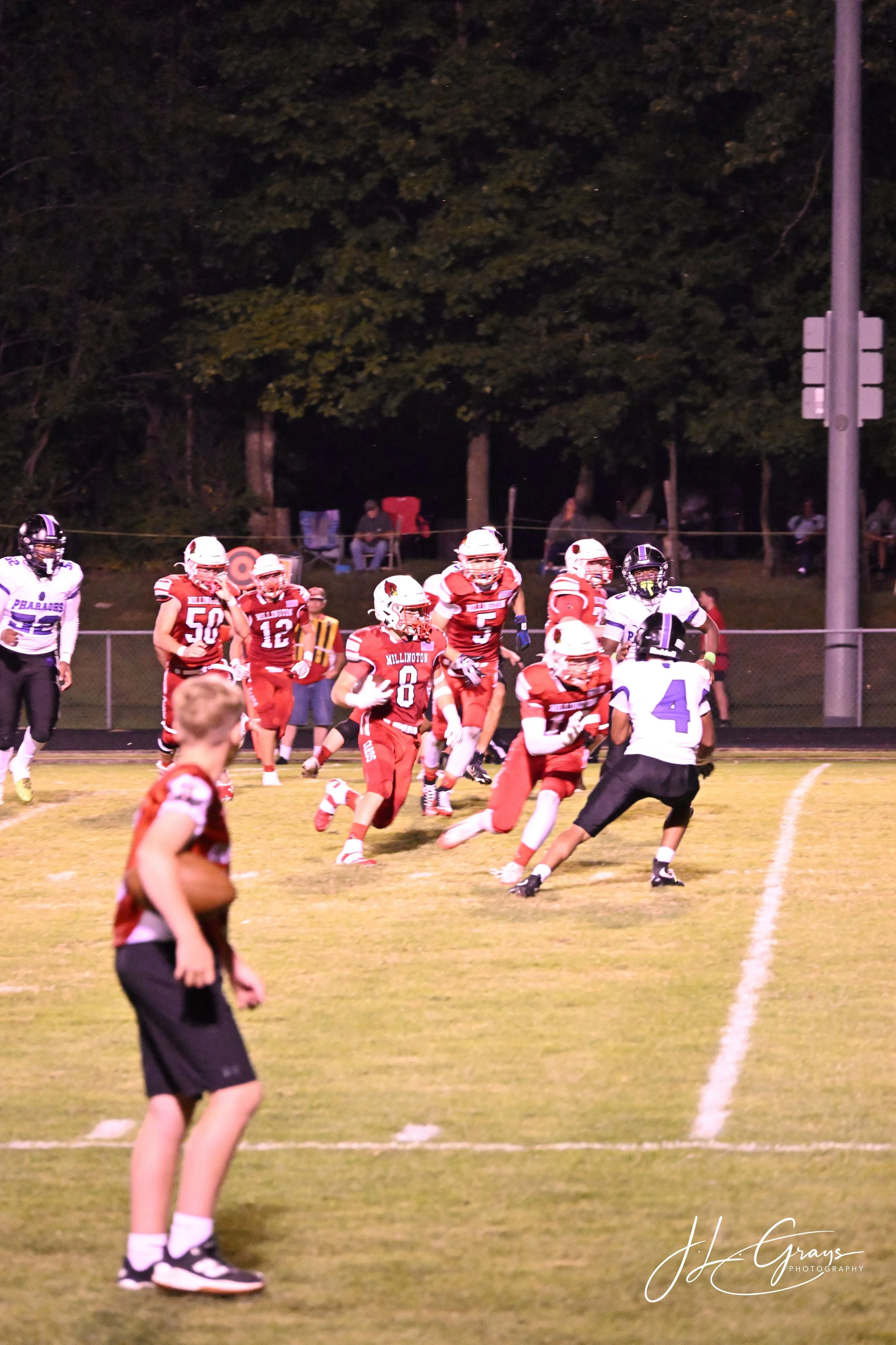 A nighttime football game with players in red and white uniforms and a player in a black and white uniform. The players are on a grassy field with trees and spectators in the background.