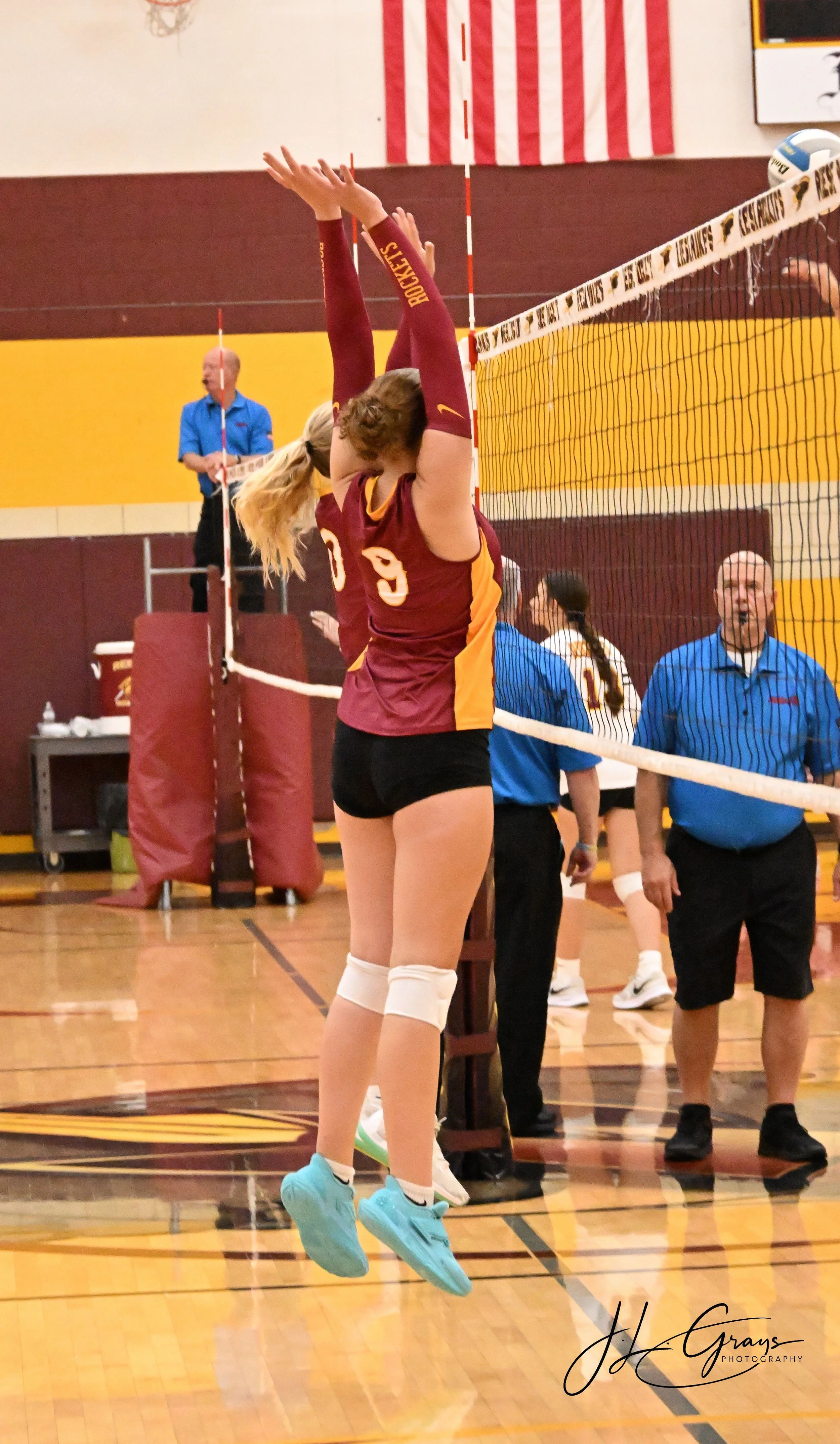 A female volleyball player in a maroon and yellow uniform with the number 9 jumps up to block the ball at the net during a game in a gymnasium. Other players, officials, and referees are visible in the background.