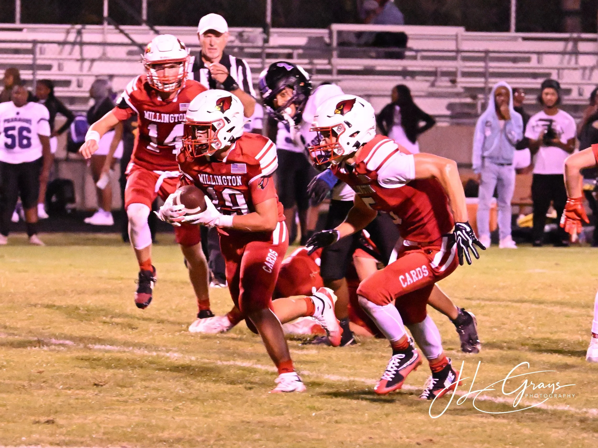 An American football game with players in red and black uniforms. One player is running with the ball, while others are blocking or preparing to make a play on the field. An official is in the background.