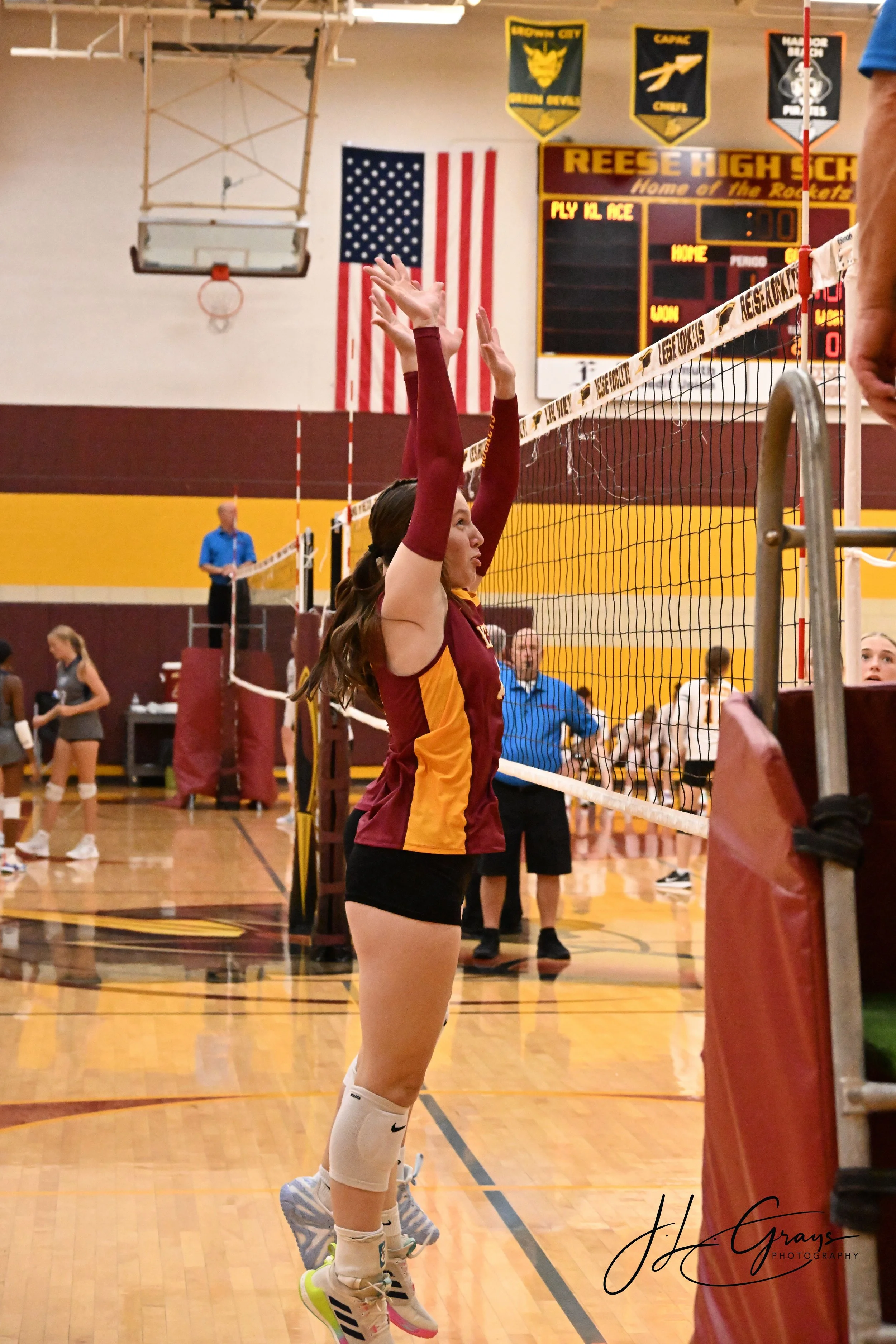 A female volleyball player in a maroon and yellow uniform jumping to block the ball at a volleyball net in a gymnasium. An American flag and school banners hang on the wall in the background.