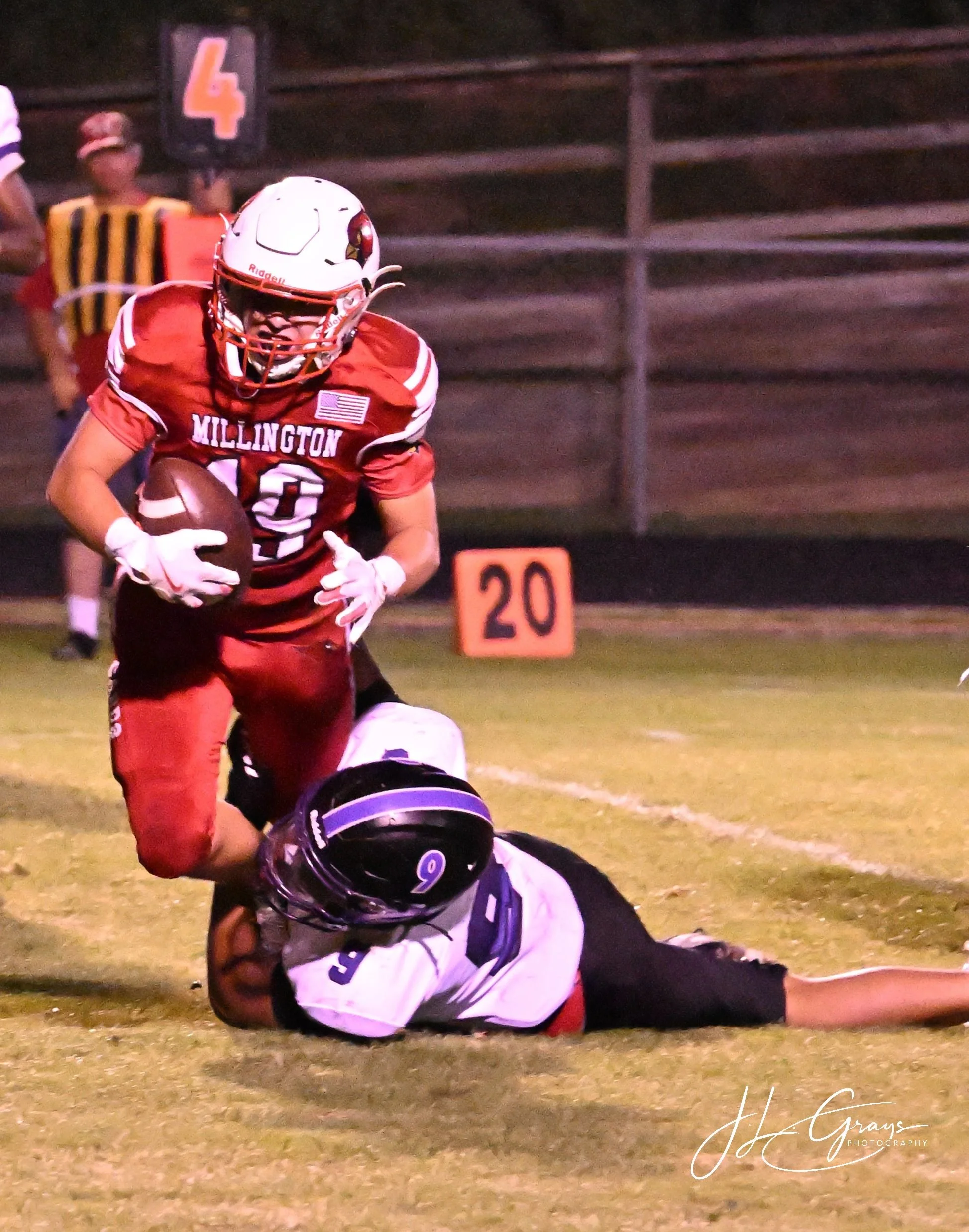 An American football game with players on the field. A player in a red uniform with the word 'Millington' is running, while another player in a white uniform with the number 9 is on the ground. A referee in the background holds a yard marker with the