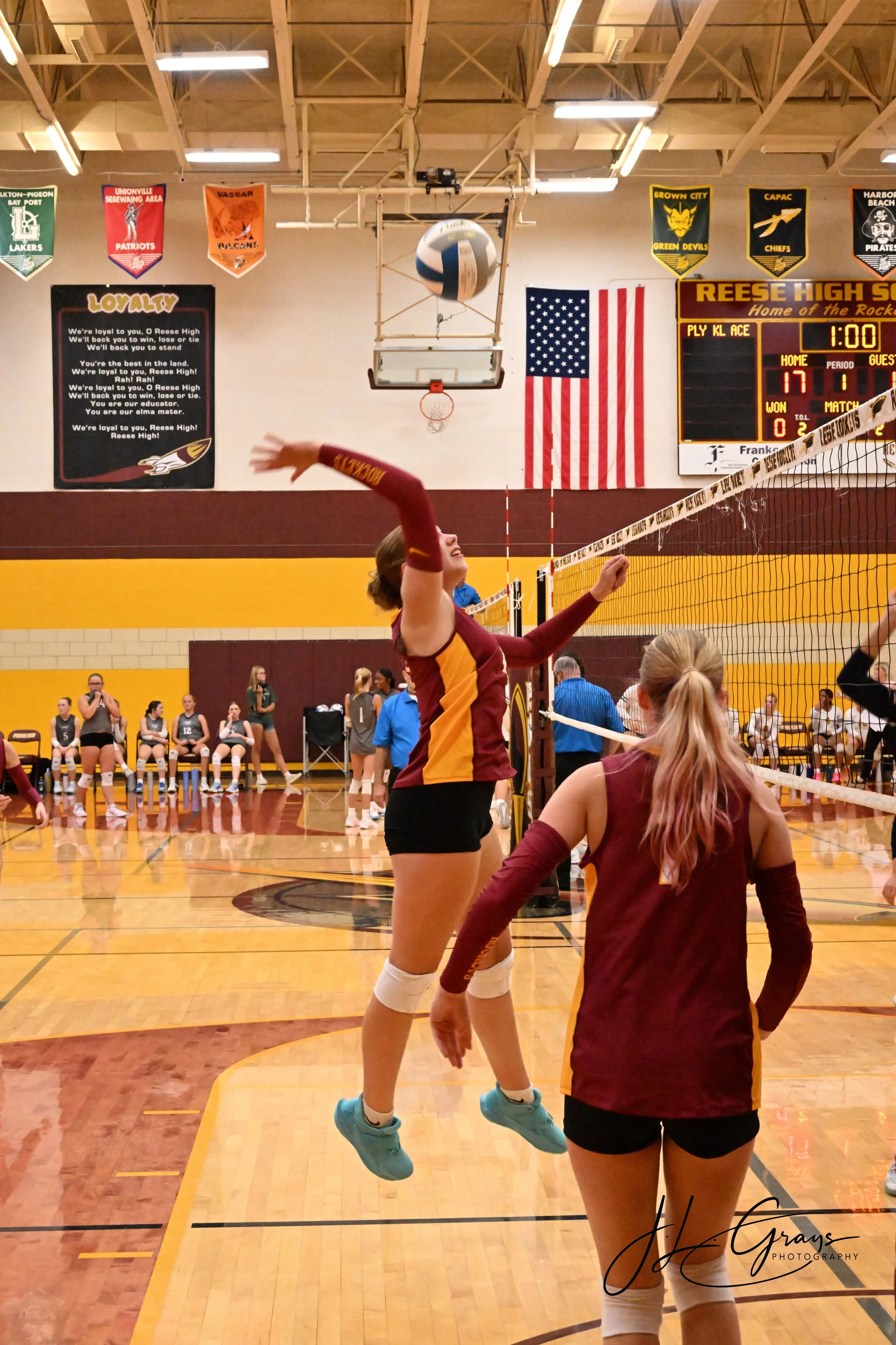 A volleyball game in progress at Reese High School gymnasium with players warming up or practicing. The gym features a gym scoreboard, American flag, and school banners hanging from the ceiling.