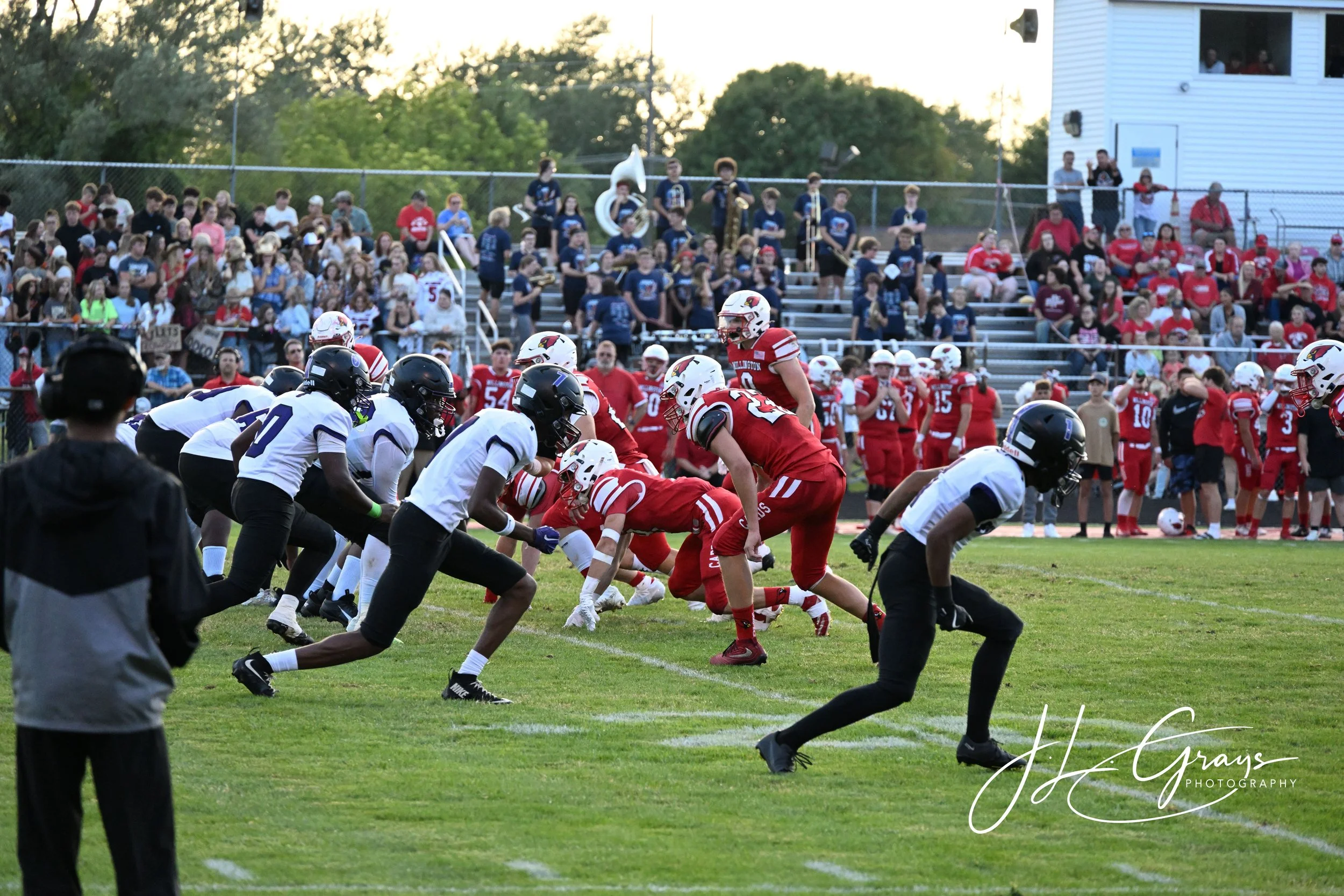 A football game in progress with players from opposing teams lining up at the line of scrimmage. Spectators are seated on bleachers in the background. The scene is set outdoors on a grassy field during the evening.