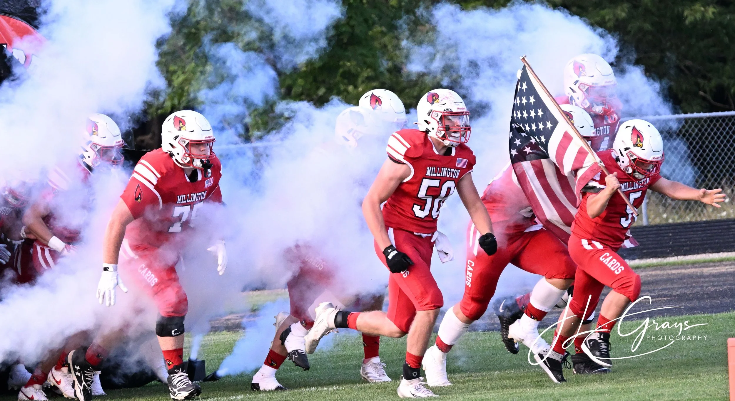 High school football team dressed in red uniforms with a cardinal mascot runs onto the field through smoke, led by a player carrying a flag, with trees in the background.