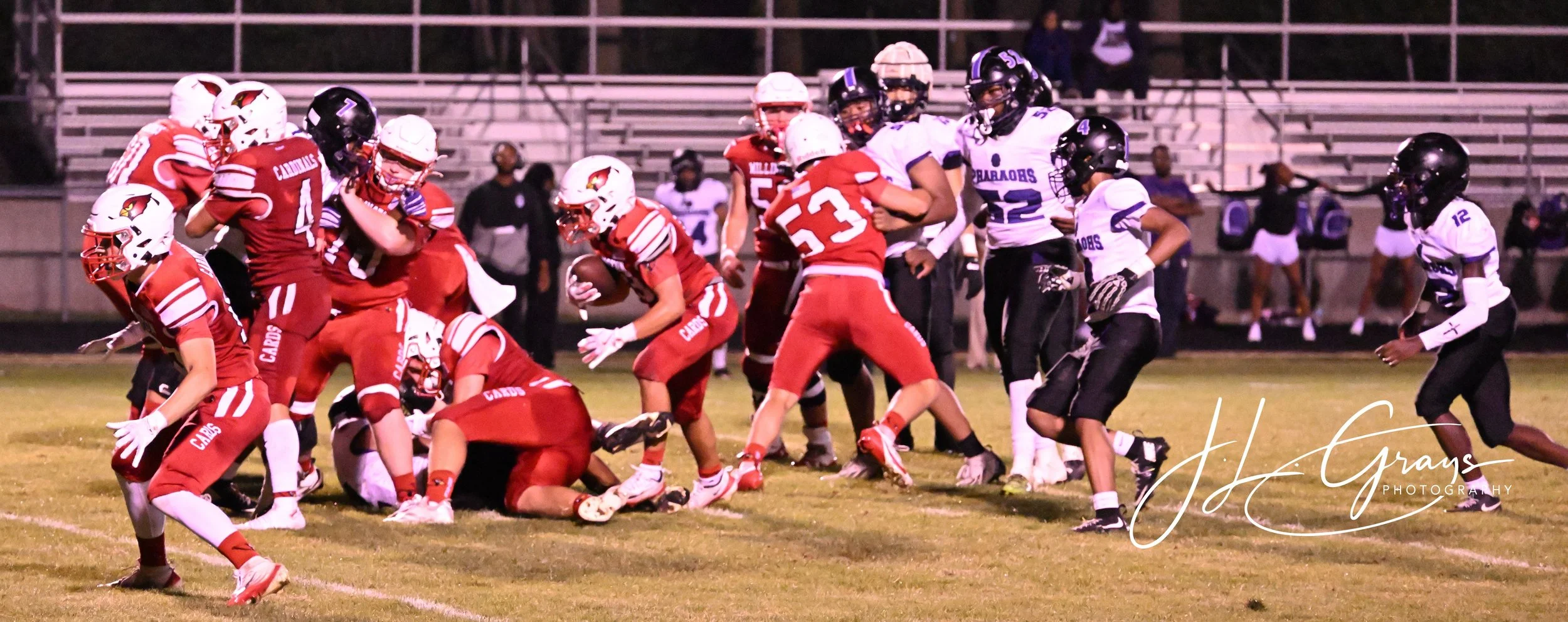 A high school football game in progress on a grass field; players in red and black uniforms are tackling and blocking players in white and purple uniforms; stadium bleachers are visible in the background; night setting with stadium lights.