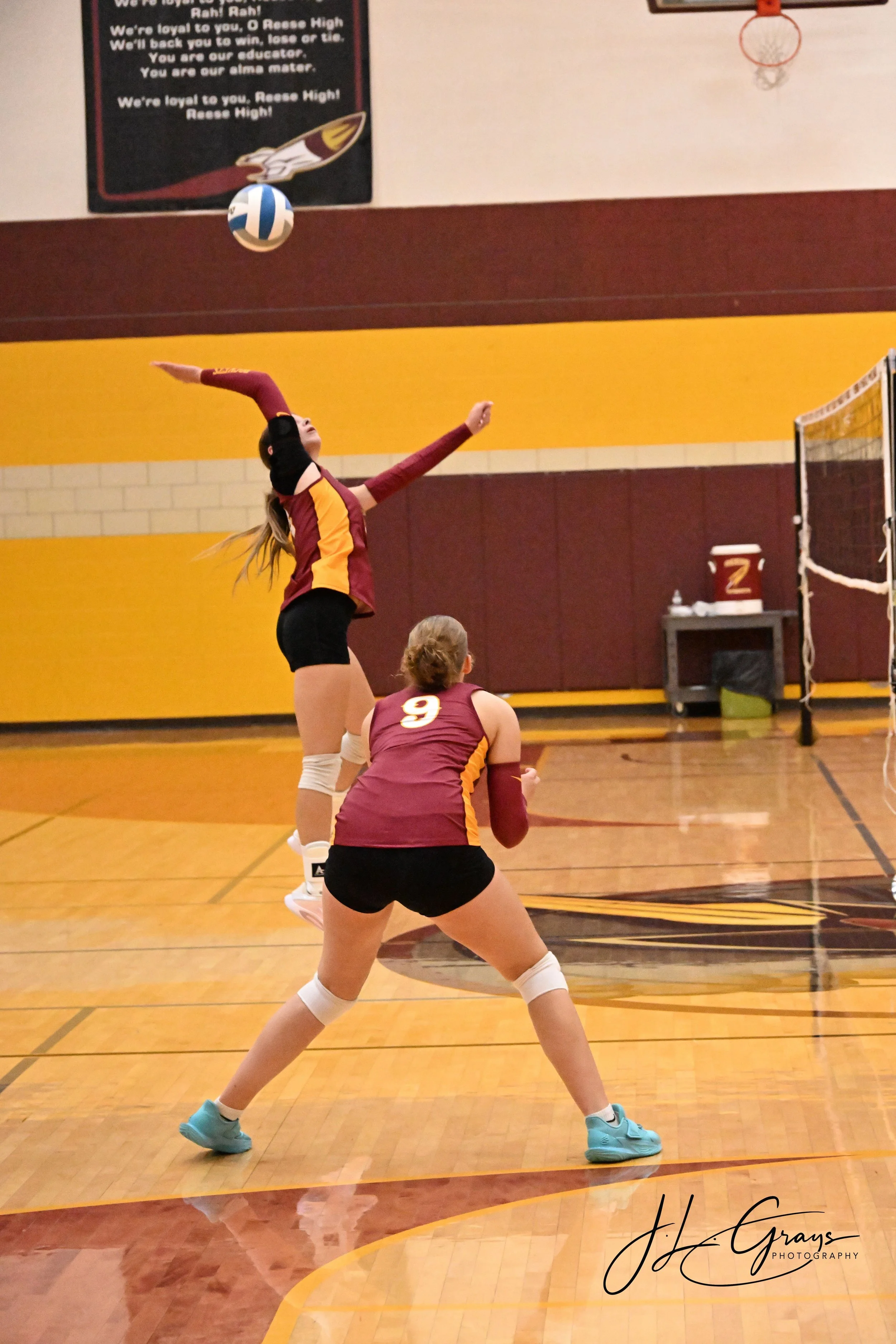 Volleyball player in maroon and yellow uniform preparing to serve on a wooden court, with a teammate in a similar uniform and a volleyball net in the background.