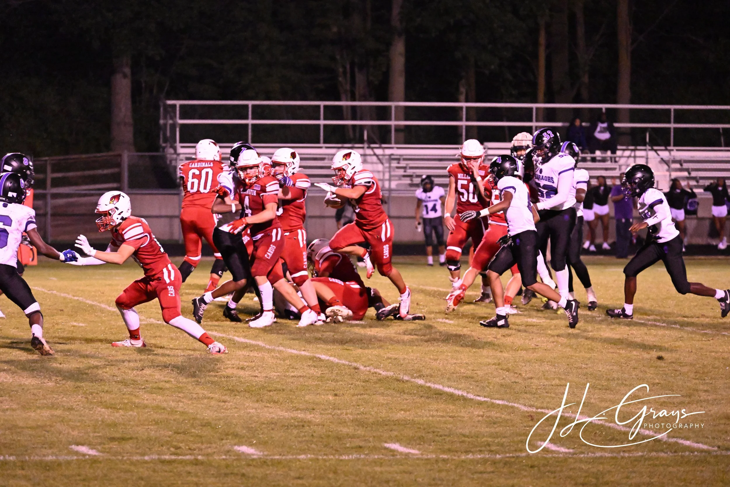 A nighttime football game with players in red and white uniforms and black helmets, and players in black and white uniforms. The players are engaged in a tackle on the field, with a few spectators and empty bleachers in the background.
