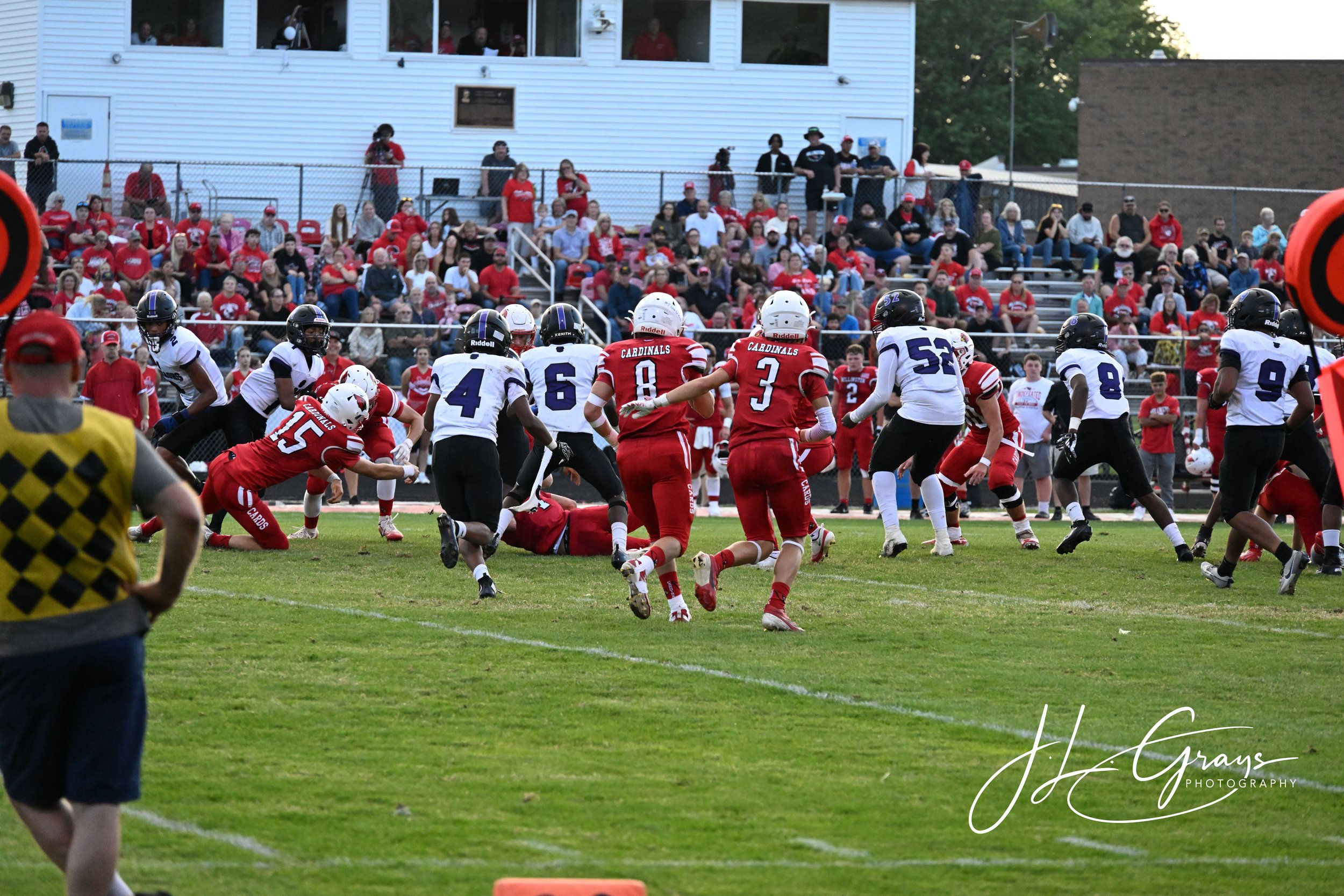 An American football game in progress with players from the Cardinals team in red uniforms and the opposing team in white and purple uniforms. The game is taking place on a grassy field with spectators in the stands and people watching from a buildin