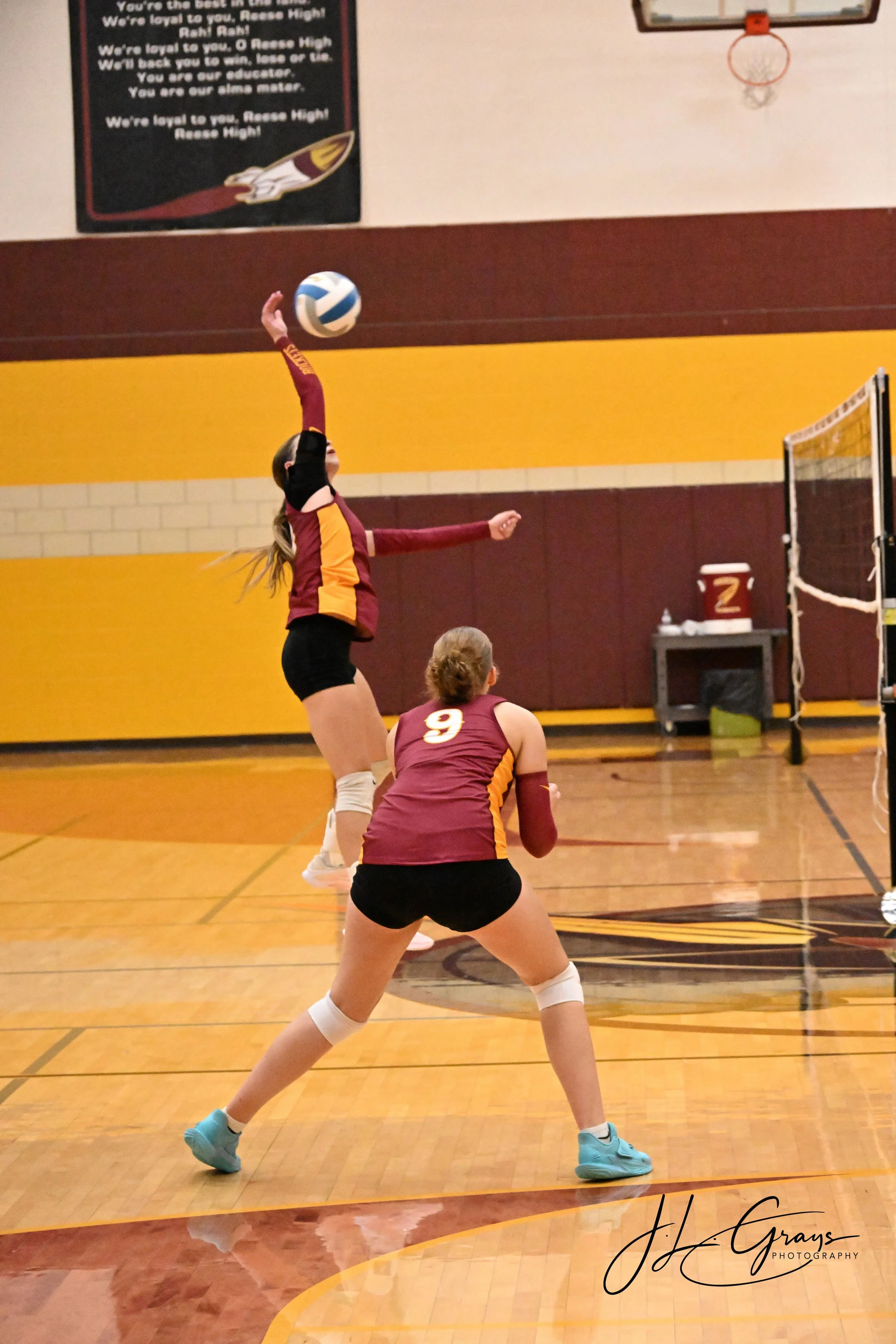 A female volleyball player in a maroon and yellow uniform jumps to hit the ball over the net during a game in a gymnasium with a hardwood floor and a basketball hoop on the wall.