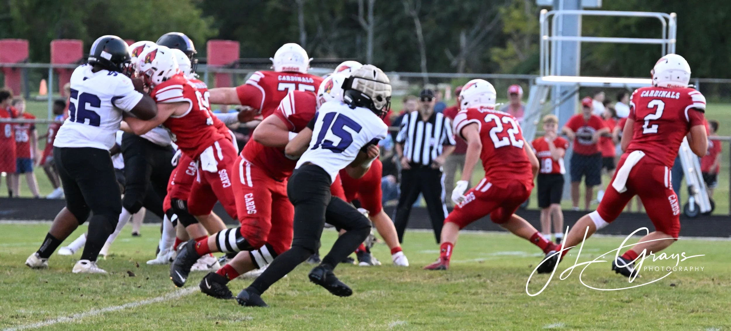 Football game with players in red and white uniforms on a grassy field, some players tackling, others running, with spectators in the background.