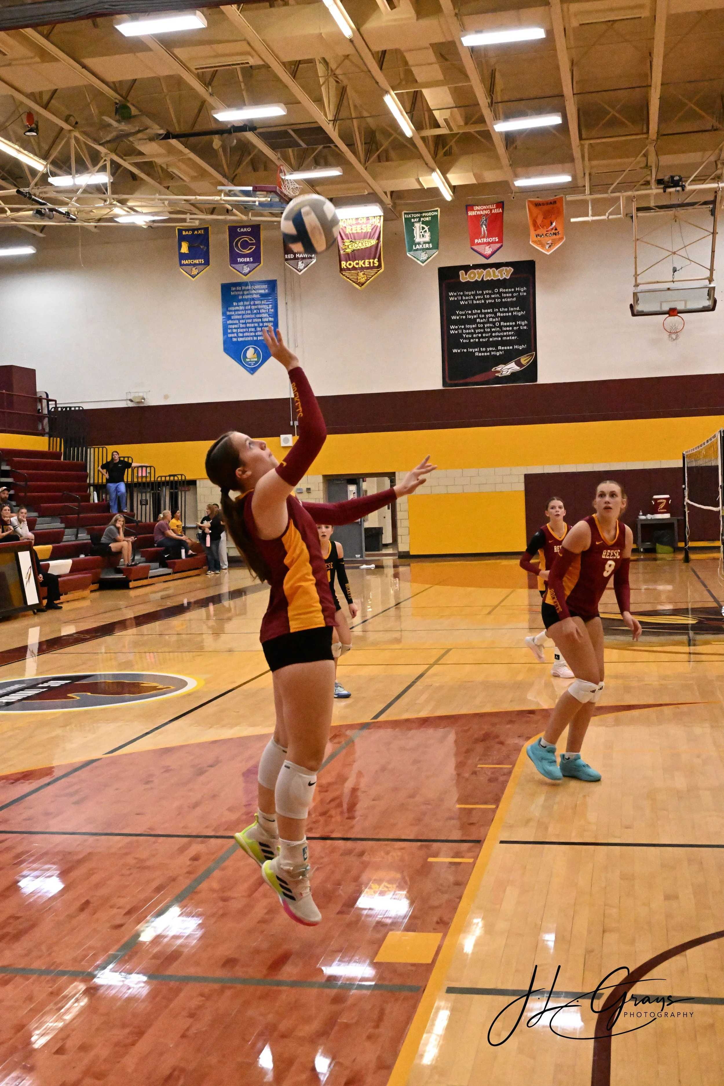 A girl jumping to hit a volleyball in a gymnasium. She is wearing a maroon and yellow sports uniform with knee-high socks and volleyball shoes. Other players are visible in the background, and there are banners hanging from the ceiling with sports te