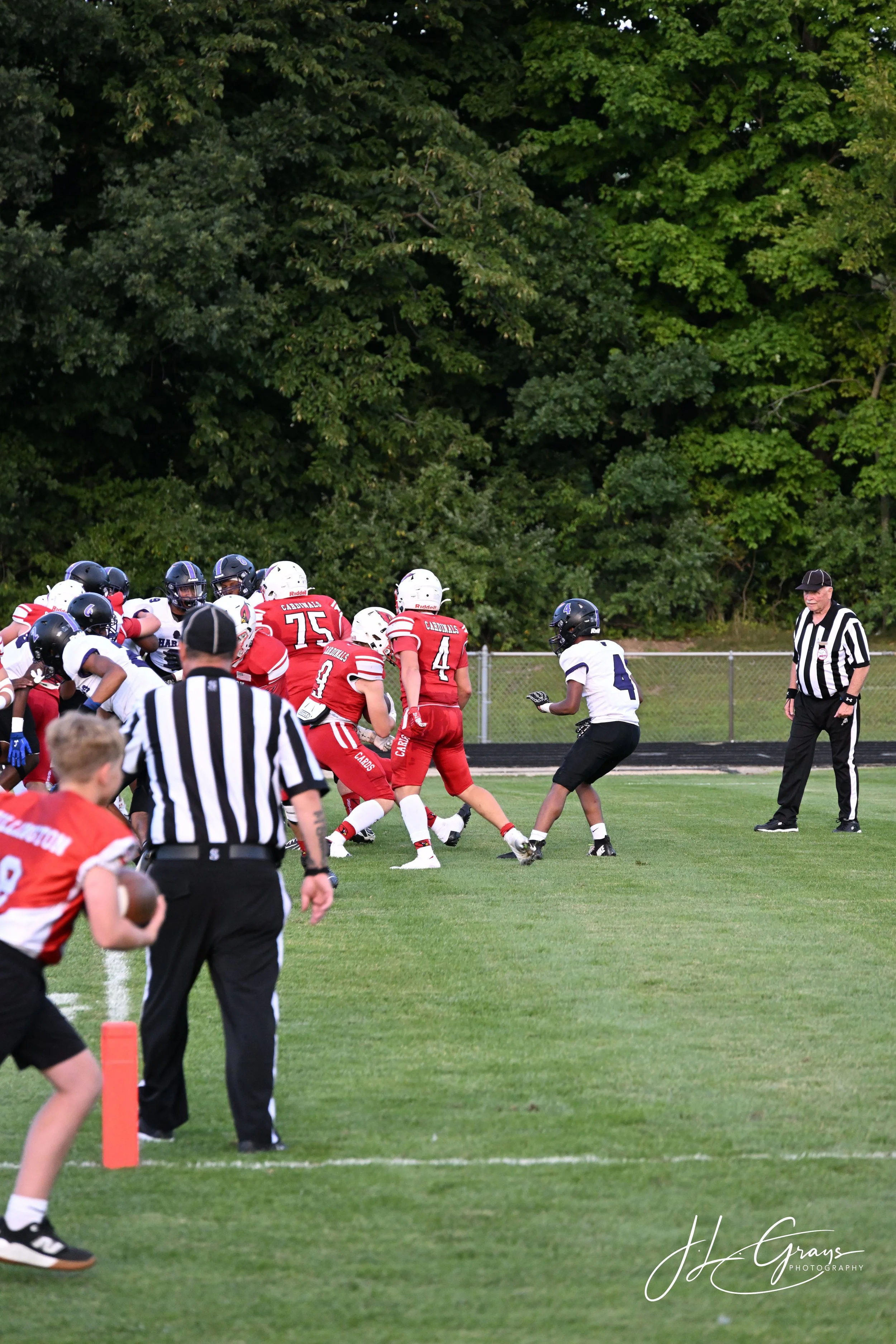 High school football game with players in red and white uniforms and referees on a grassy field with trees in the background.