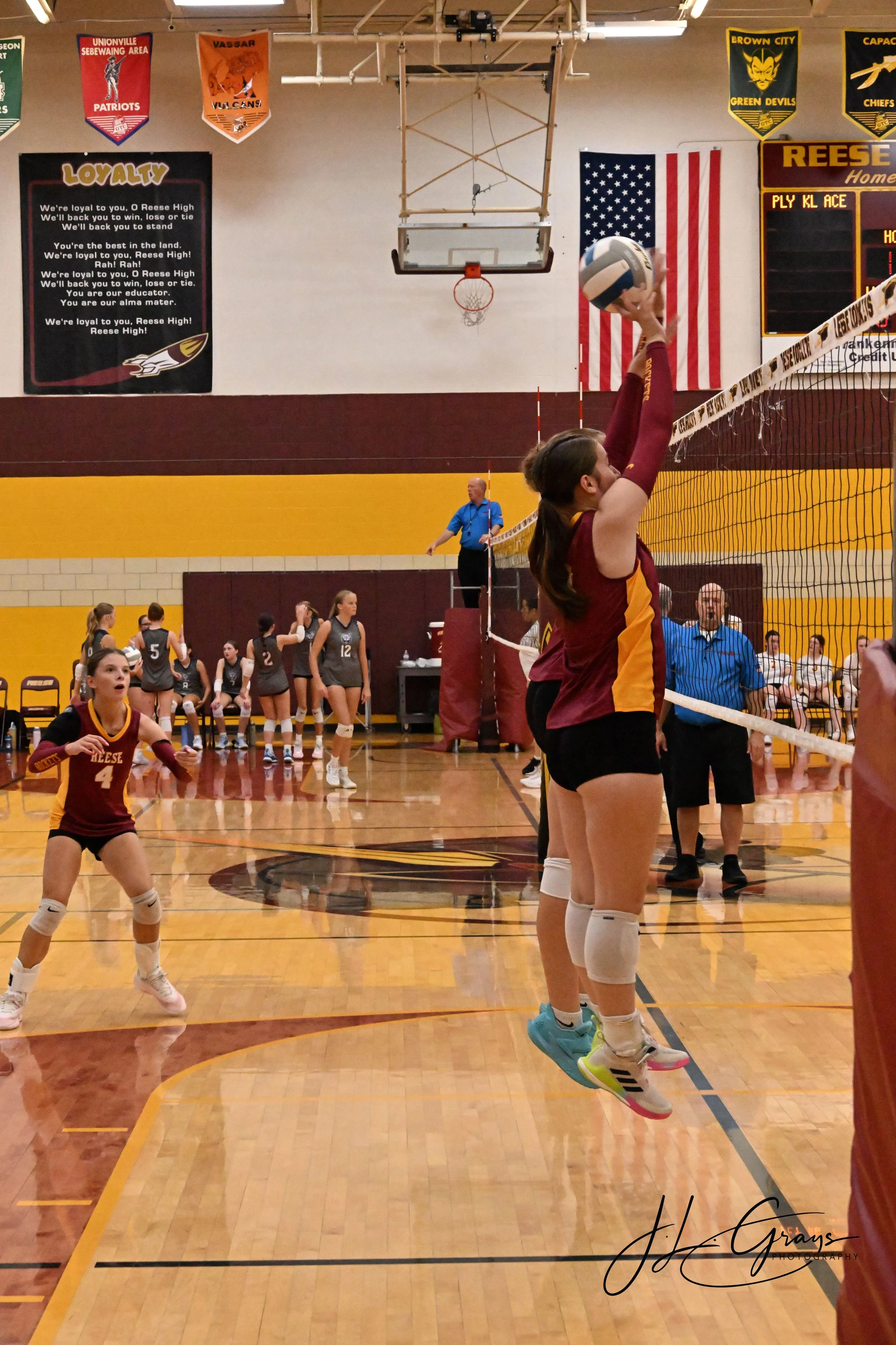 A volleyball game in progress in a gymnasium, with players warming up or practicing. One player in the foreground, wearing a maroon and yellow uniform with the number 4, is preparing to serve or hit the ball. Other players are visible in the backgrou